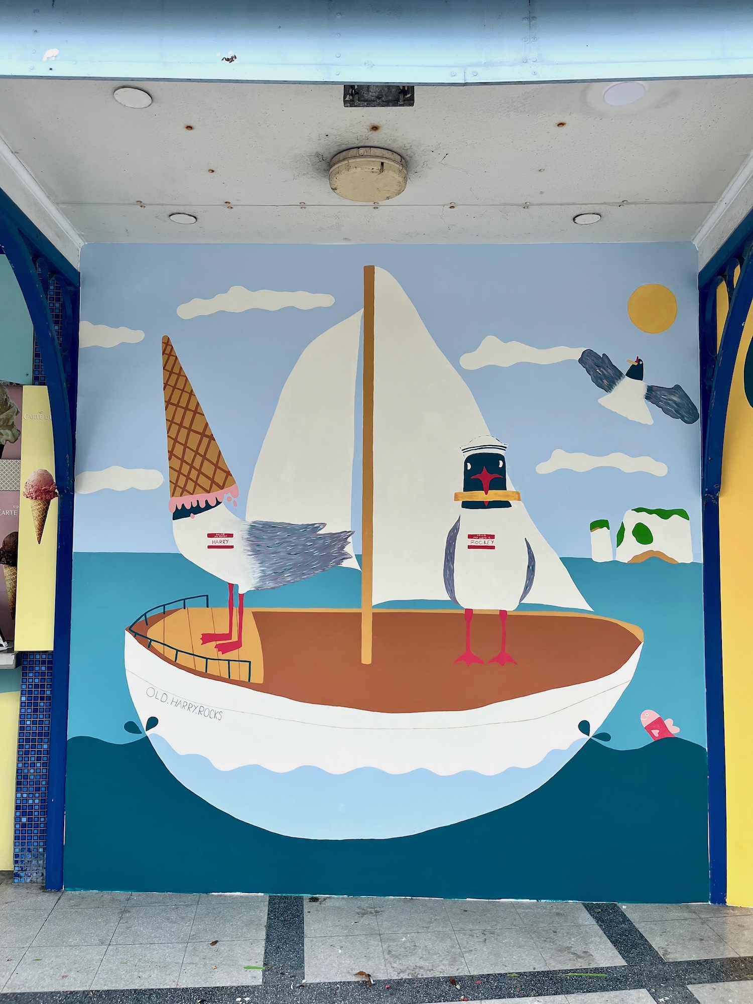 A colourful mural of two cartoon birds on a sailboat, painted on a wall beneath a ceiling light and framed by blue columns.