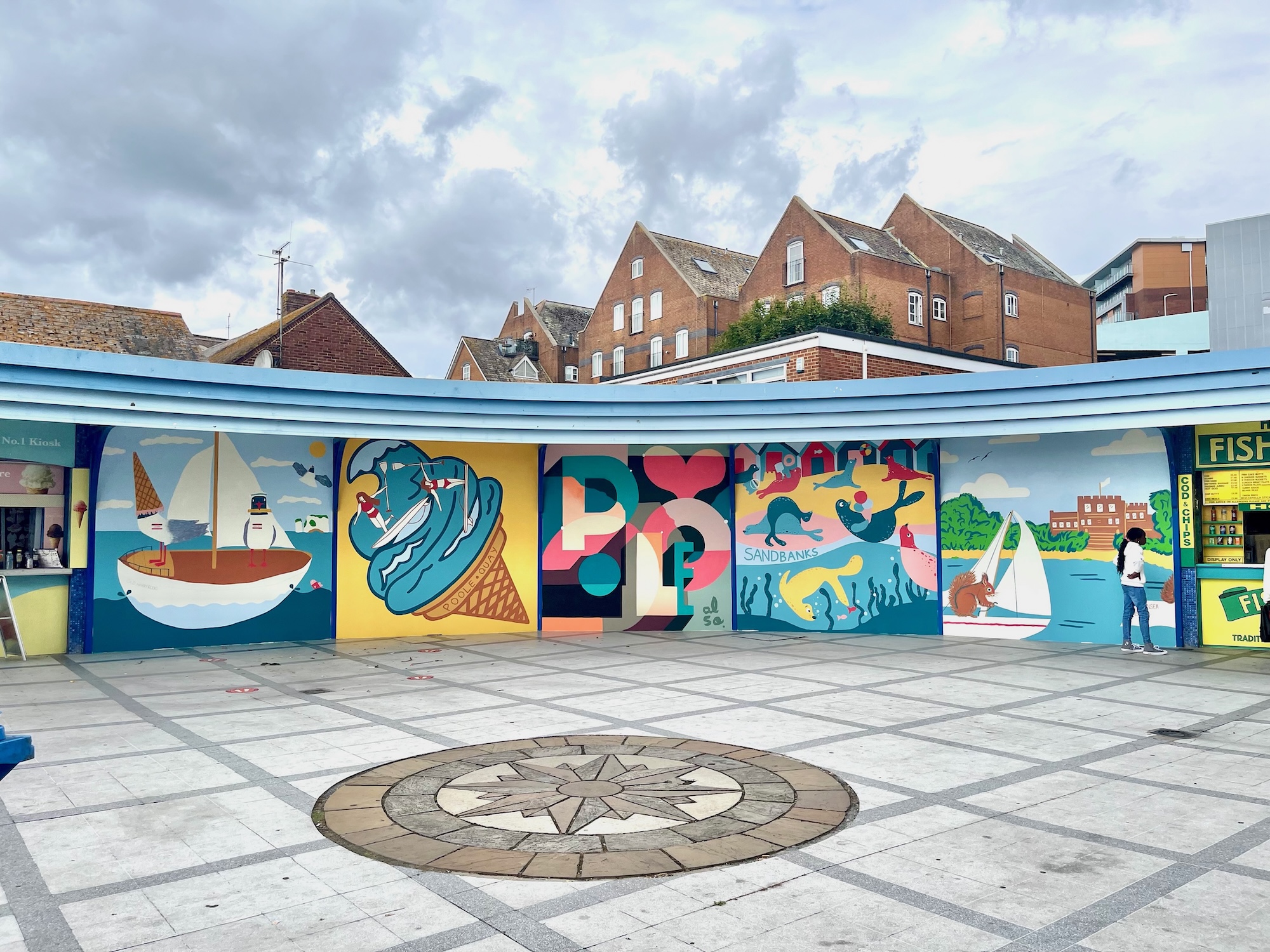 A colourful seaside mural spans a curved wall in an open paved square, with sailboats, dolphins and stylised lettering beneath brick buildings and a cloudy sky.