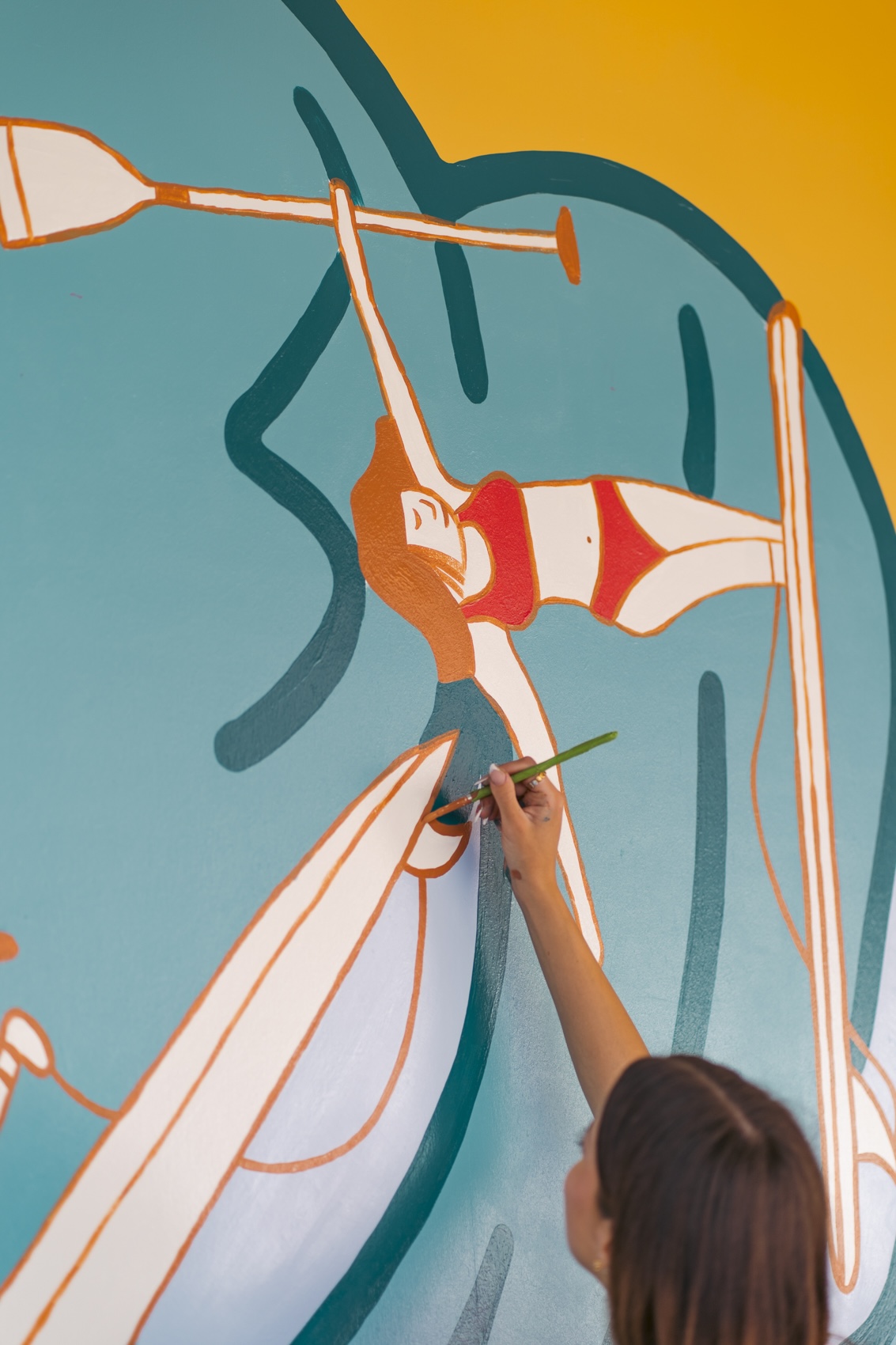 A person paints a colourful mural on a wall, adding details with a brush to a stylised figure against a blue and orange background.