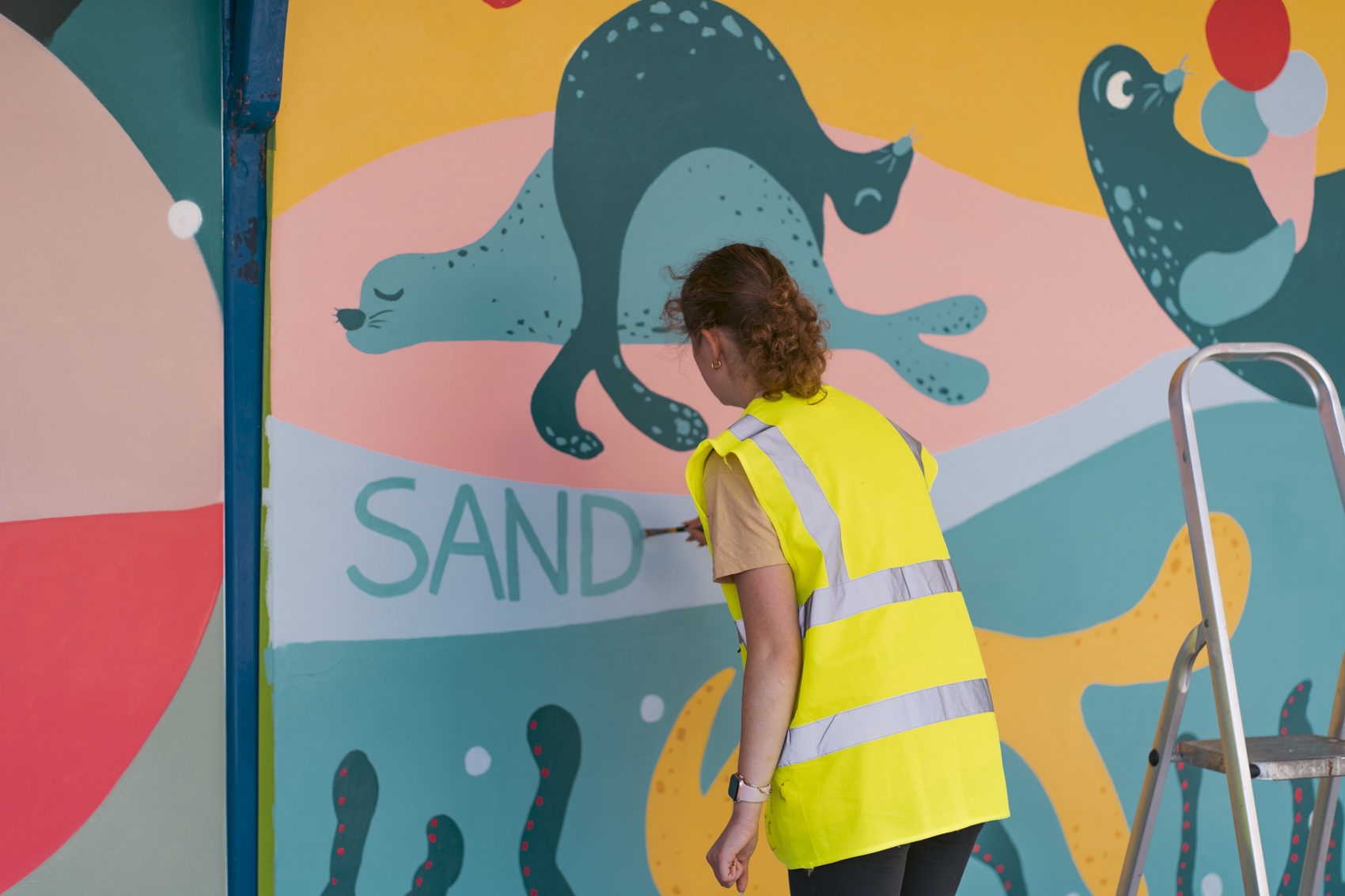A person in a high-visibility yellow vest paints a colourful mural on a wall, with a step ladder beside them and the word “SAND” visible.