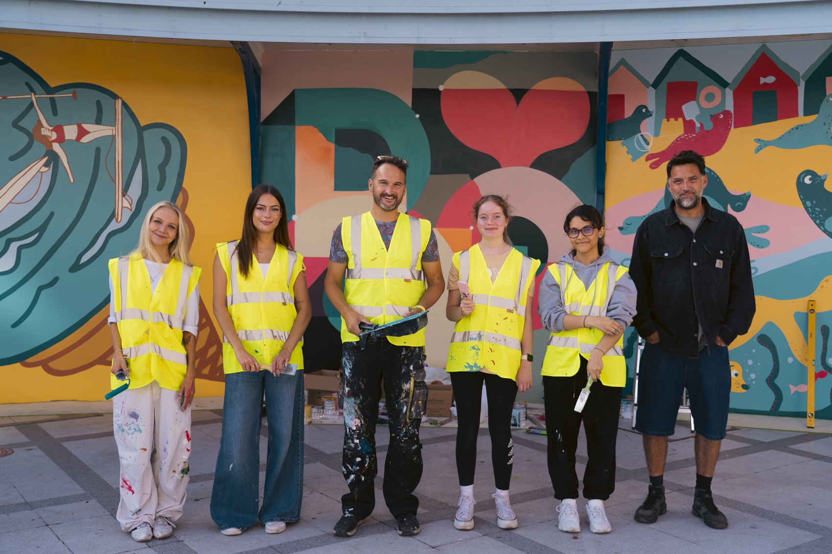 Six people stand in high-visibility yellow vests in front of a colourful mural wall, with the central man holding paint tools.