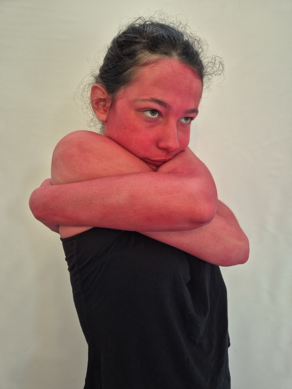 Woman with arms crossed, skin and arms painted bright red, wearing black top against pale grey background.