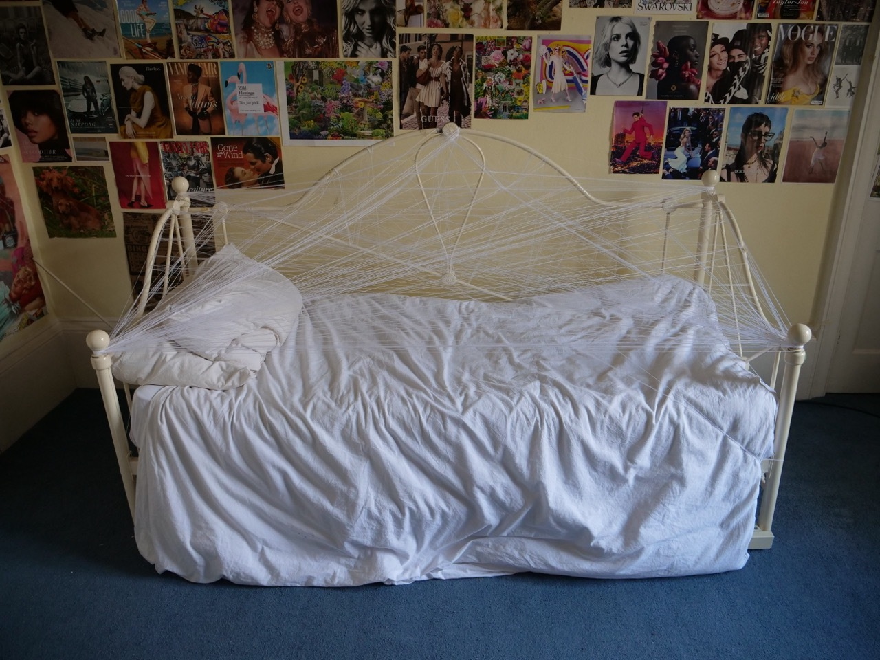 White metal daybed with rumpled white bedding and webbed canopy. Wall covered in colourful posters and magazine clippings. Blue floor.