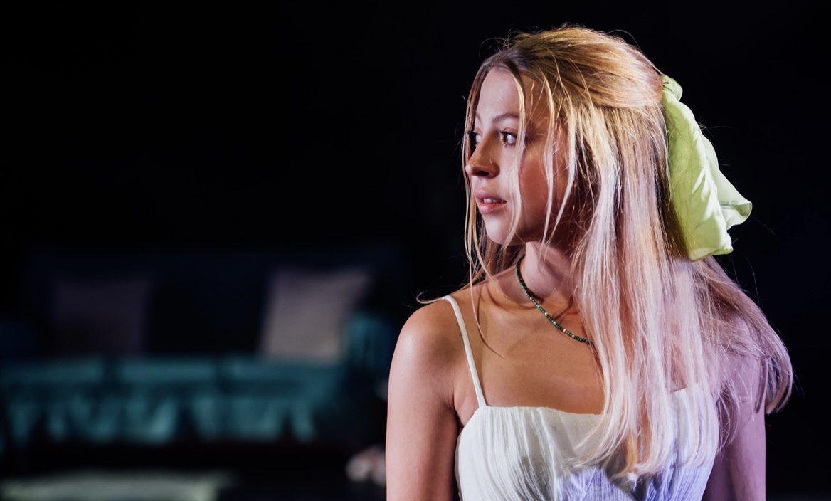 A close-up photo of a dancer in profile she has long blonde hair as well as a green ribbon in her hair, she is looking over her right shoulder.