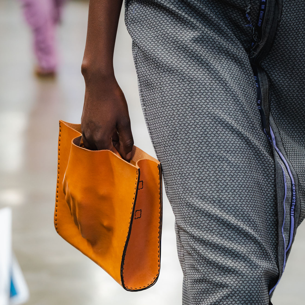 Person's hand holding bright orange leather handbag with the raised outline of the back of a hand, black stitching, wearing grey patterned trousers.