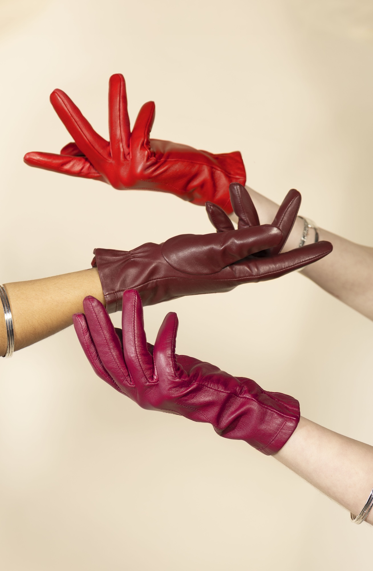 Three hands wearing red, burgundy and pink leather gloves are stacked together against a plain beige background.