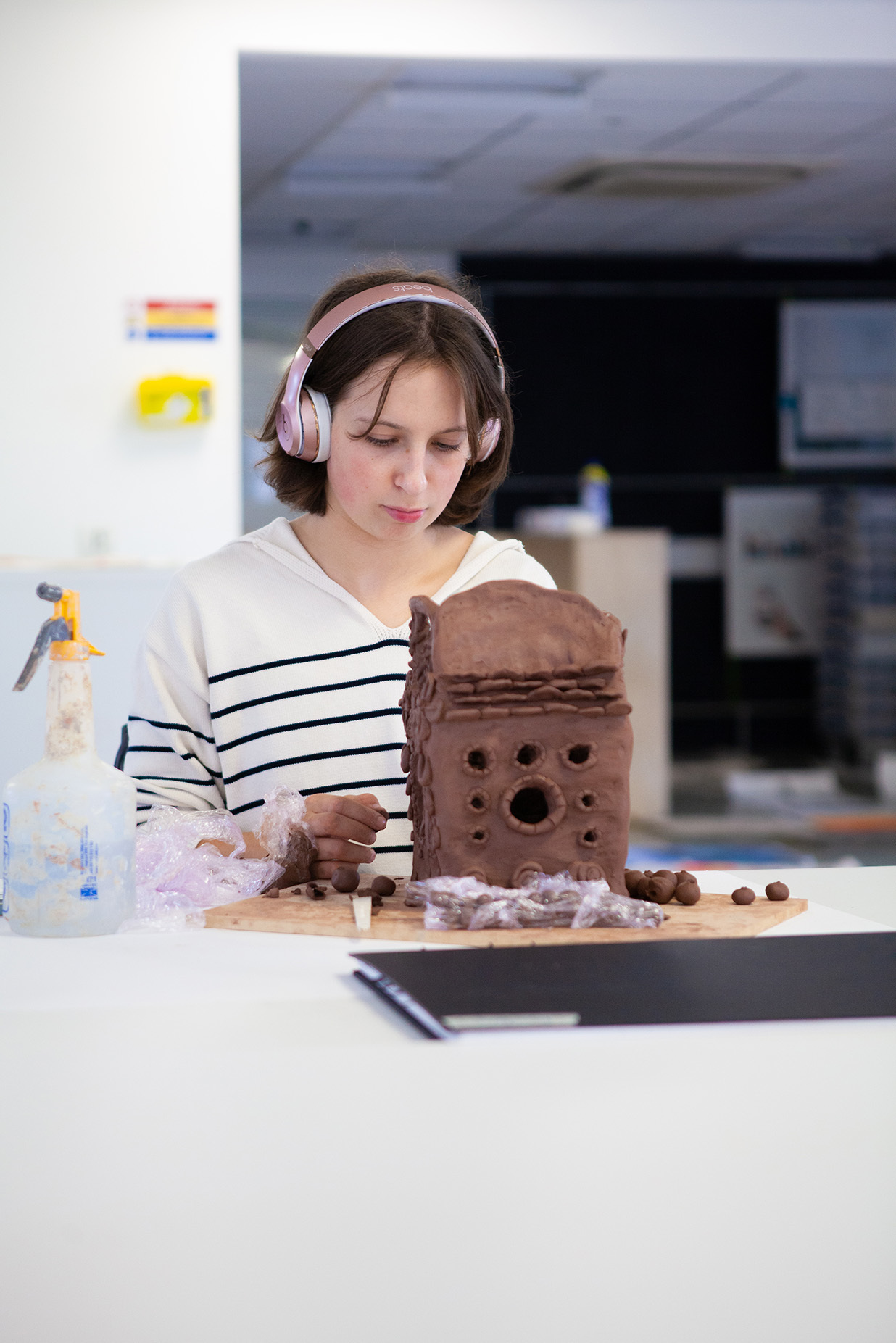 A student crafting a house out of brown clay wearing headphones.