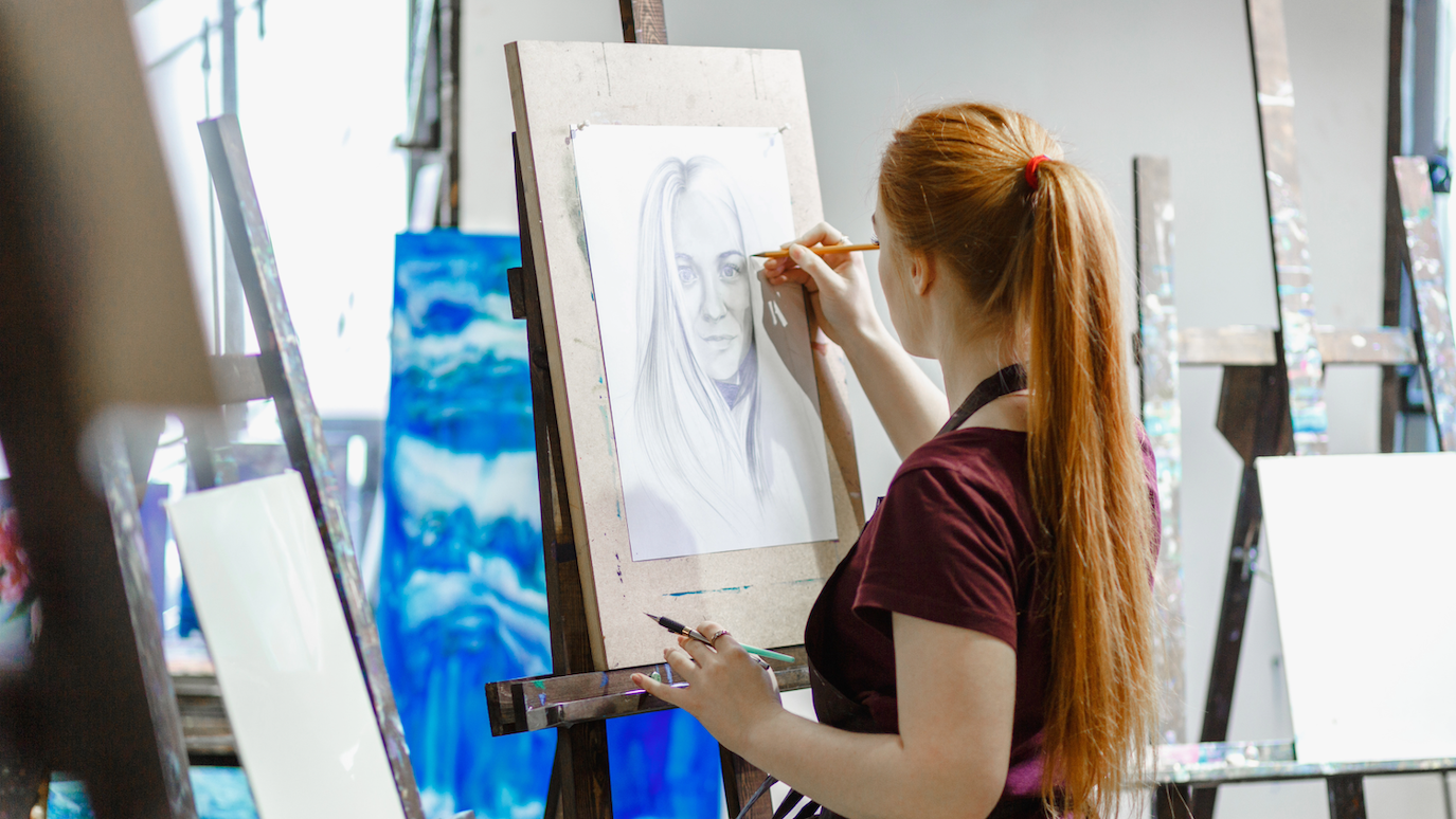 Woman with long blonde ponytail in burgundy top drawing at easel in art studio with blue painting visible in background