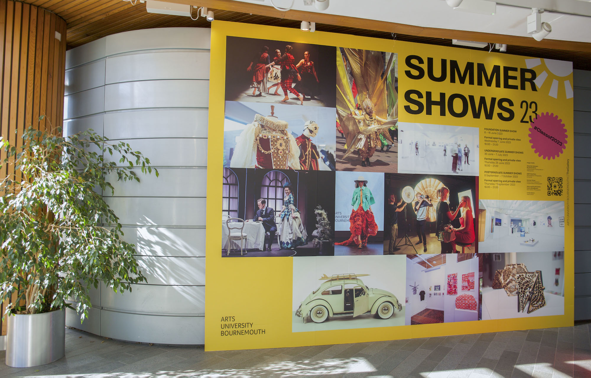 A large landscape poster with a collage of images from the AUB 2023 Summer Shows on it. The poster is standing outside a building on AUB Campus.