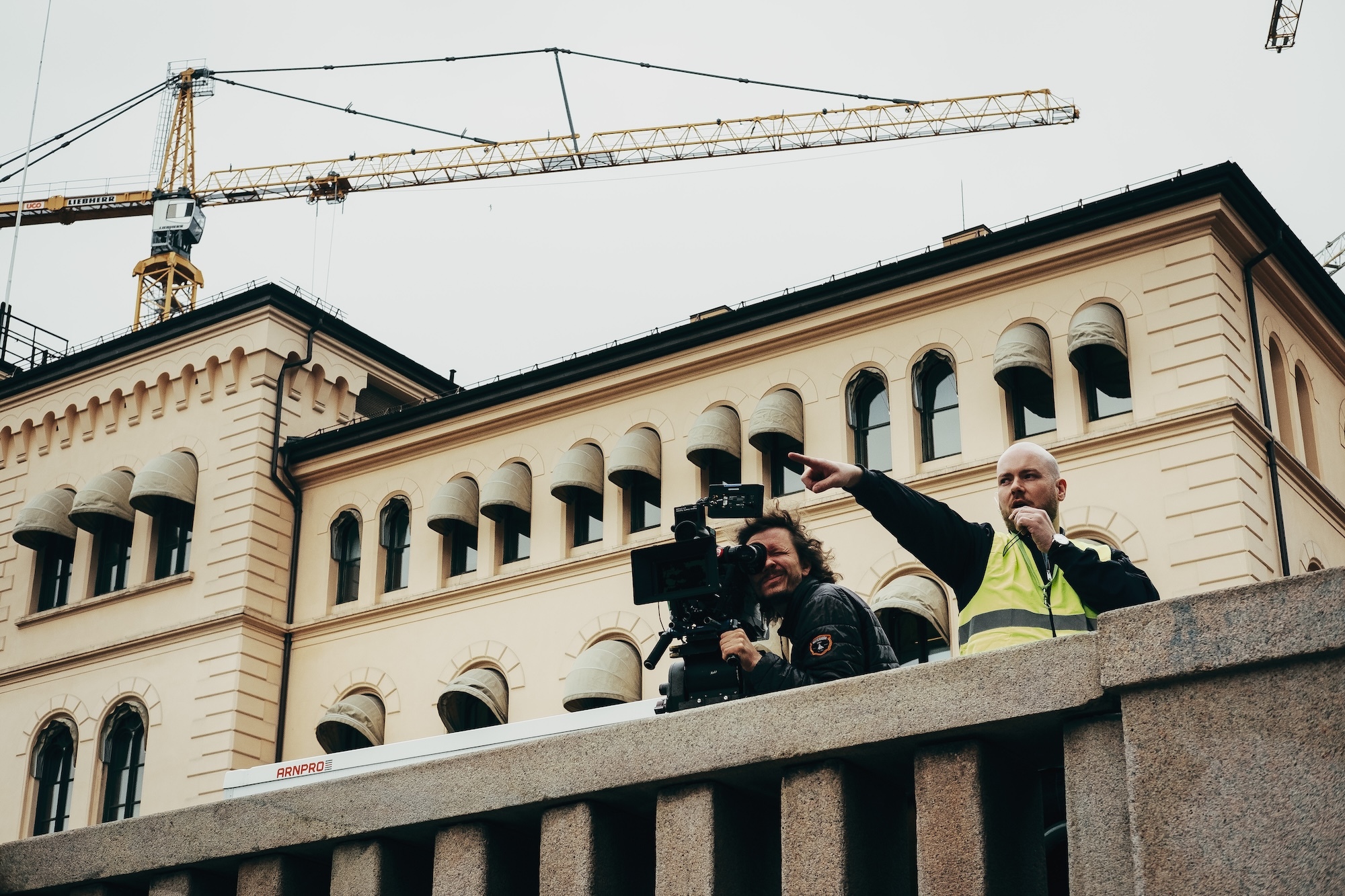 J E H Fredriksen and L T Skare on concrete balcony of cream-coloured stone building with arched windows. Yellow crane visible above structure.