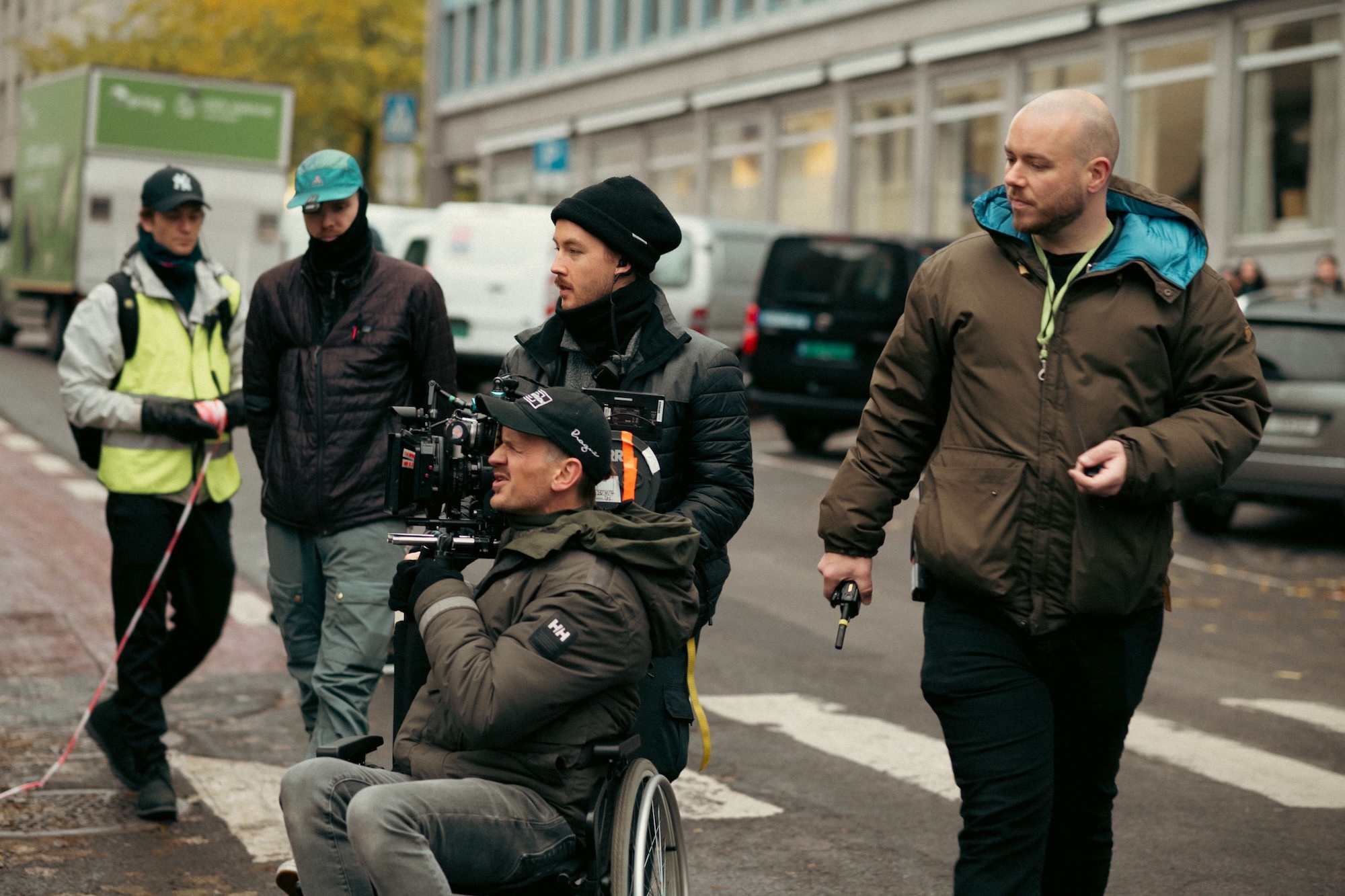 Film crew on street with camera operator seated, director standing nearby, and crew members in high-vis jackets by white building.