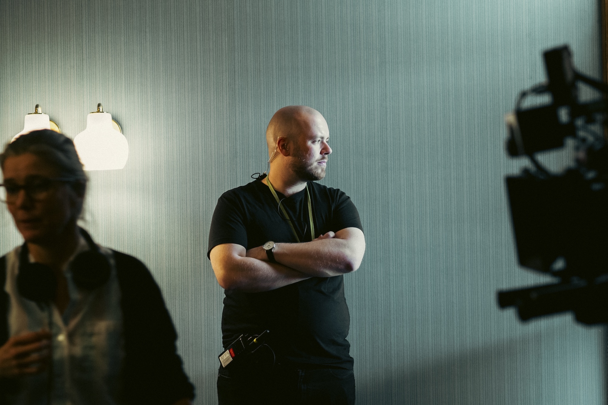 L T Skare with crossed arms on film set, camera equipment visible, bright lights in background, crew member on left.