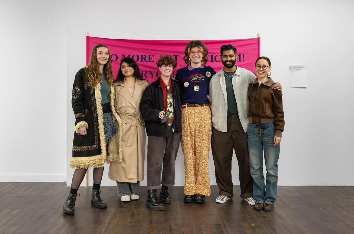 Six people standing together in front of a bright pink banner on a white wall, wearing various casual and formal clothing.