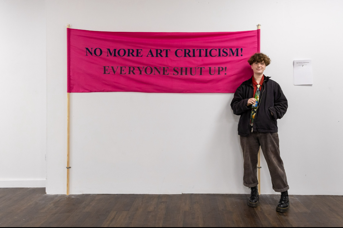 Person in dark clothing stands beside bright pink banner reading "NO MORE ART CRITICISM! EVERYONE SHUT UP!" on white gallery wall.