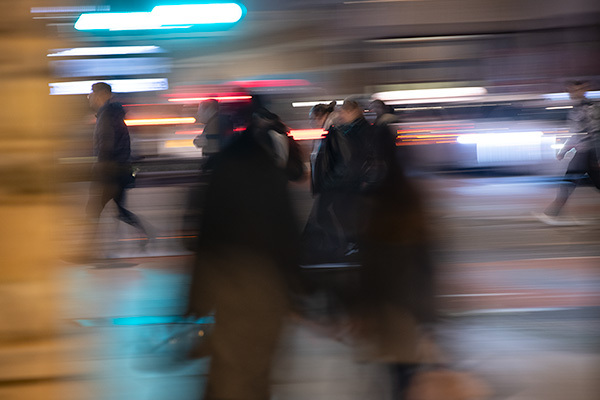 Blurred motion image showing people walking on street at night with streaked coloured lights in blue, red, and white tones.