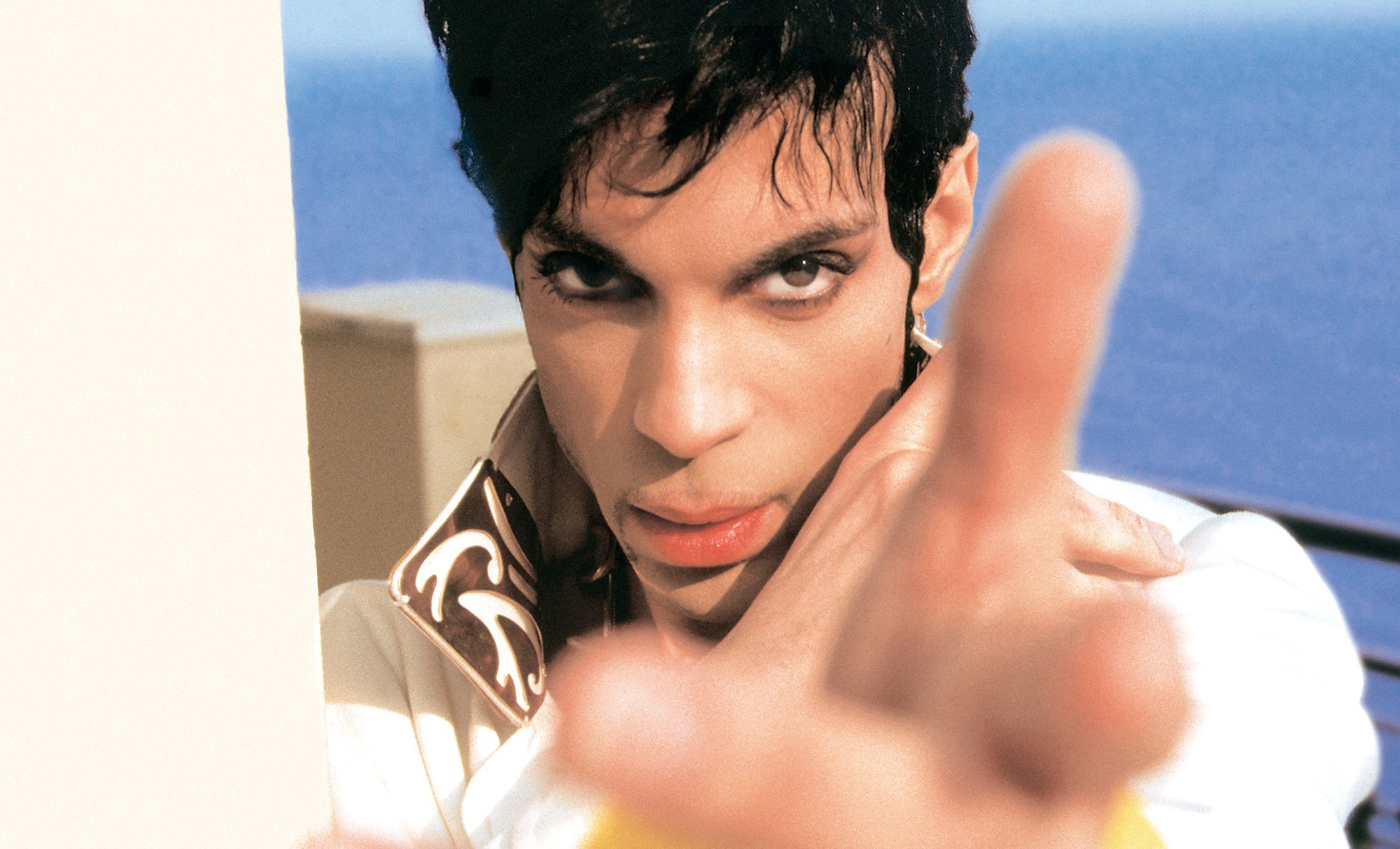 Person with dark curly hair making peace sign gesture with hand extended towards camera, wearing white shirt with patterned collar.