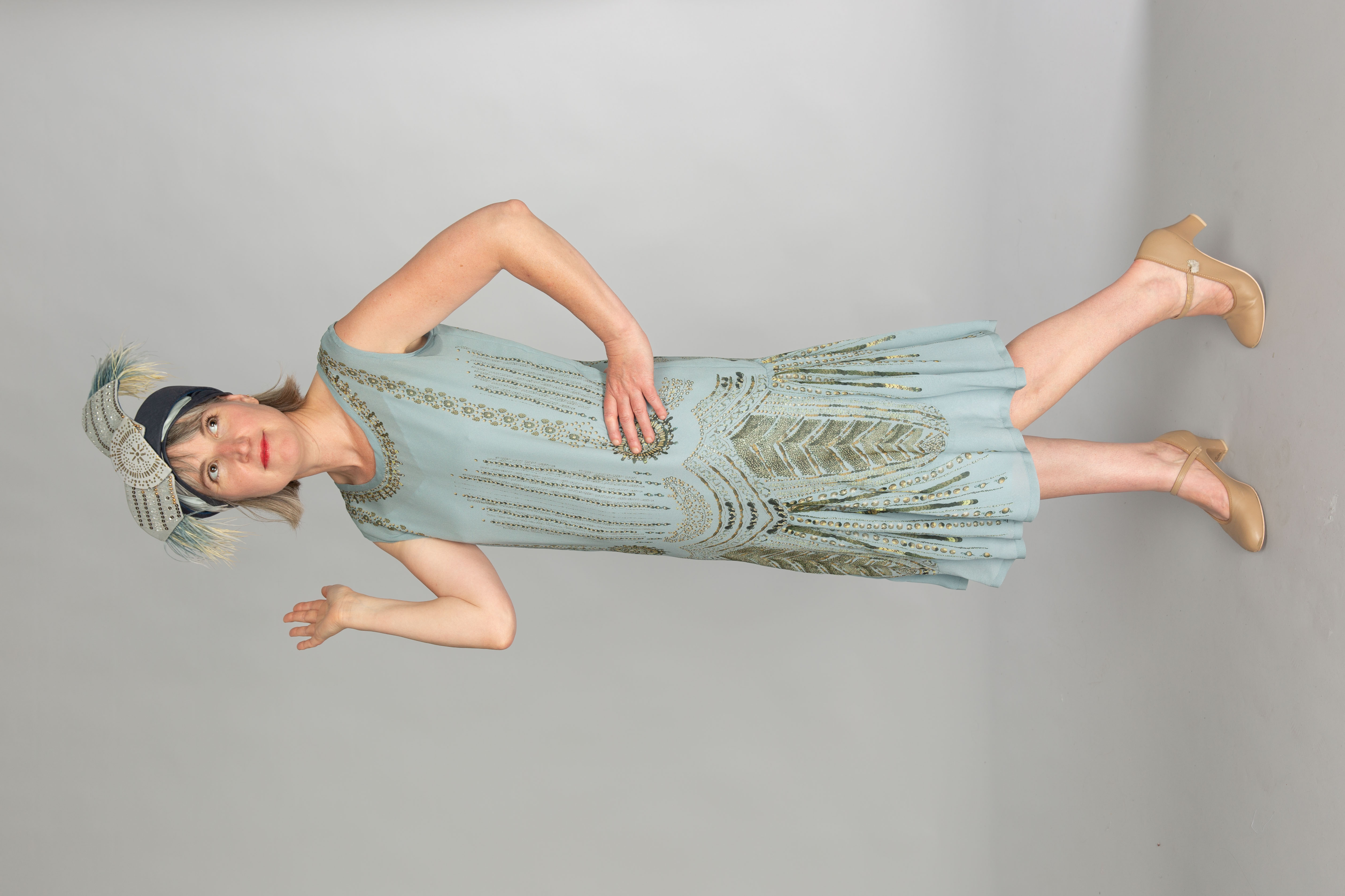 Model stood in a 1920s beaded dress replica with cape and headdress, light blue and embroidered with gold patterning with a feathered headdress.