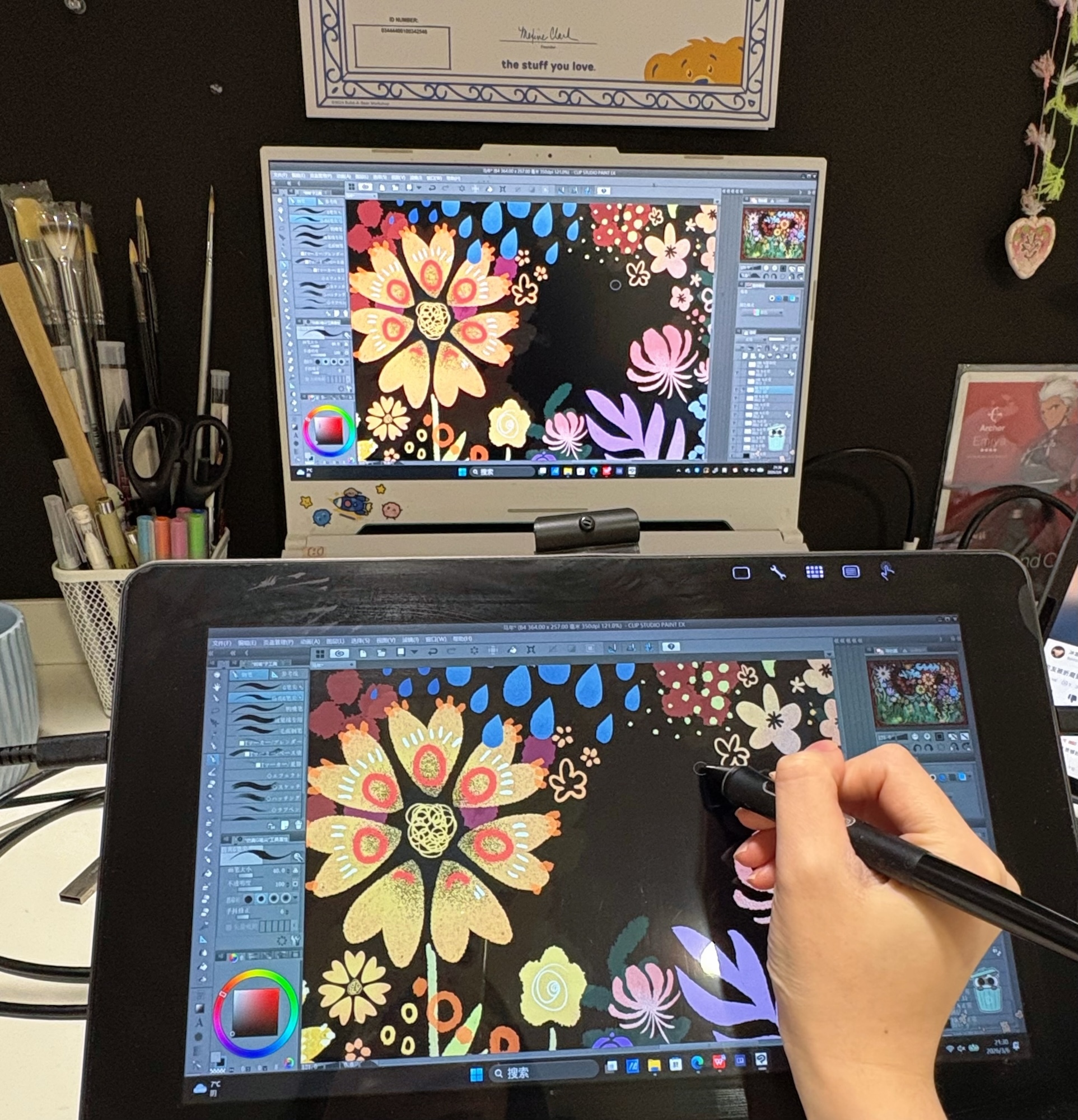 Hand holding stylus on graphics tablet displaying colourful floral digital artwork, with matching design on computer monitor behind.
