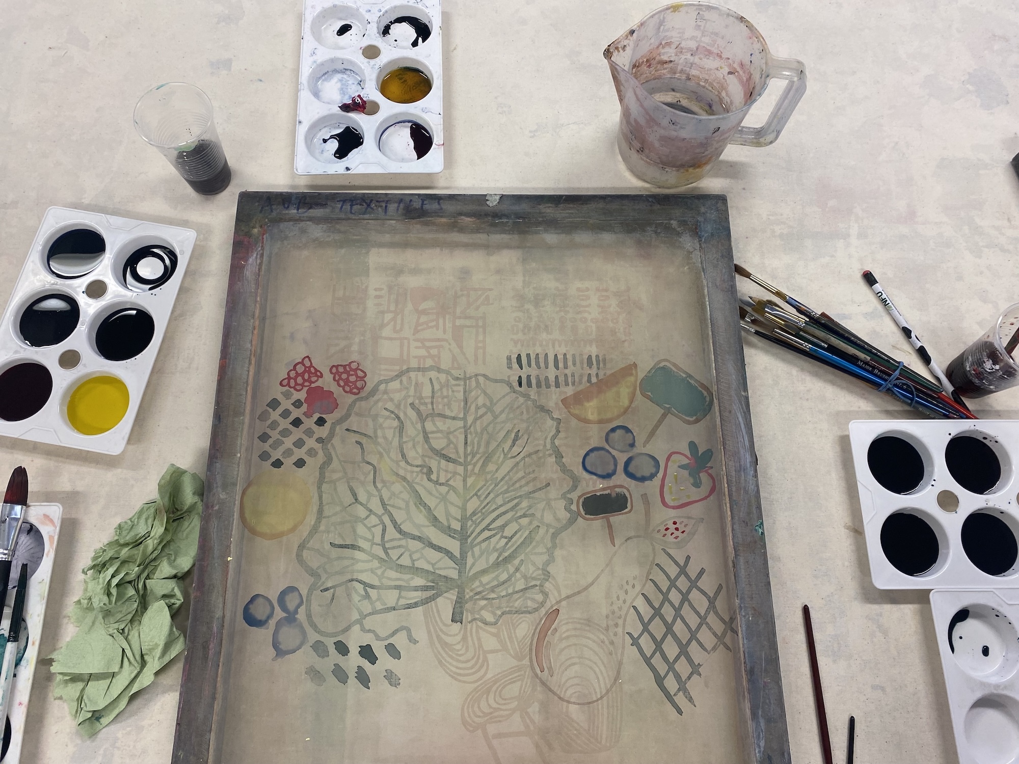 Art workspace with watercolour palettes, brushes, and glass jar surrounding central drawing board containing botanical sketches and geometric patterns in muted colours.