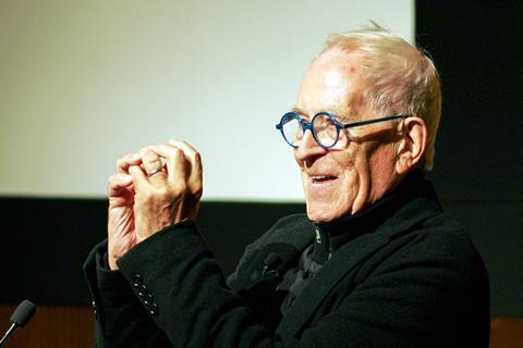 Elderly man with glasses and grey hair gesturing with raised hand whilst speaking at podium, wearing dark jacket.