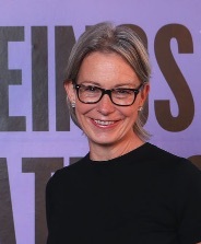 Woman with short blonde hair and black-rimmed glasses wearing dark top, smiling against purple background with partial text visible.