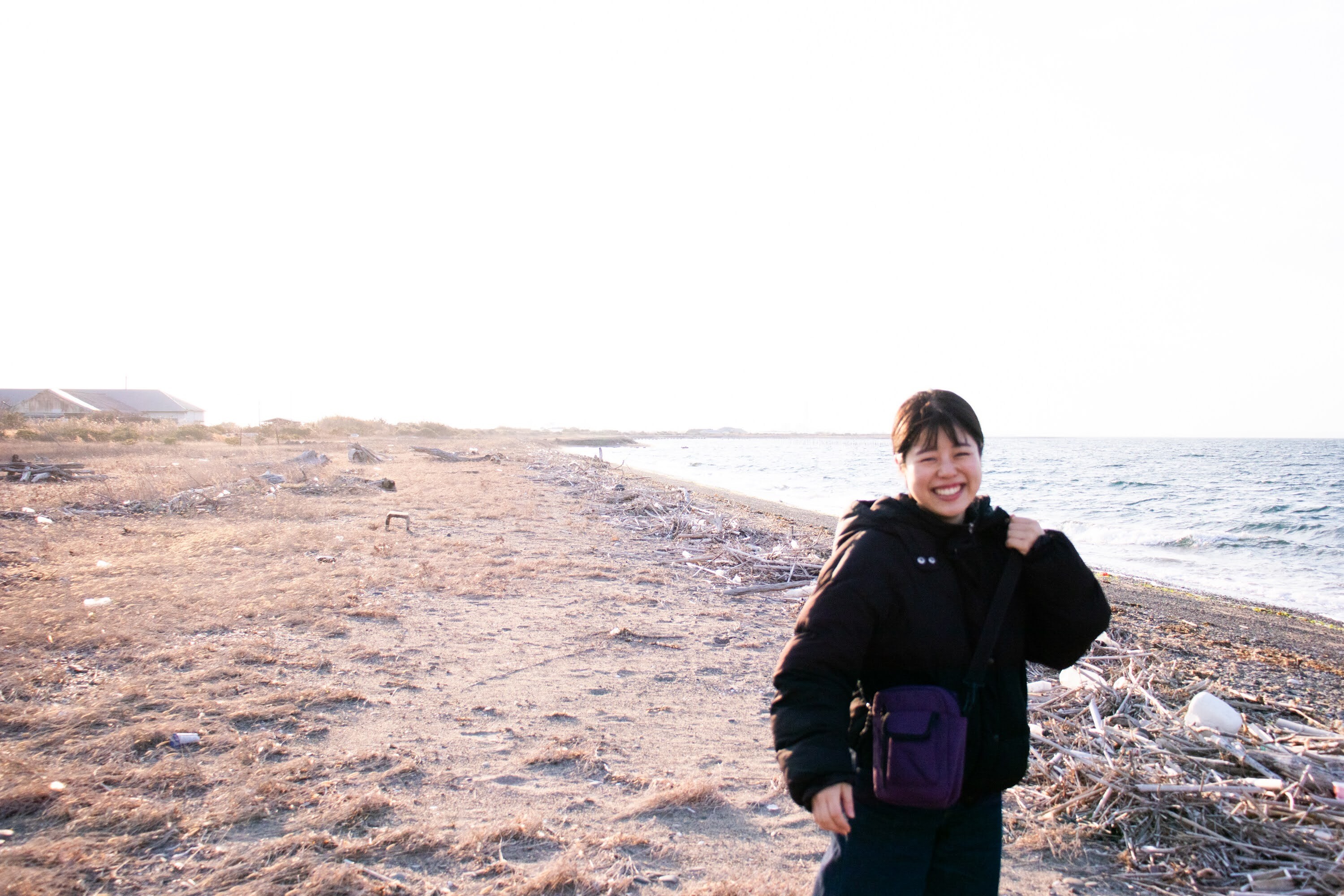 M Kadowaki in black jacket standing on rocky beach with water and distant cliffs under bright sky.