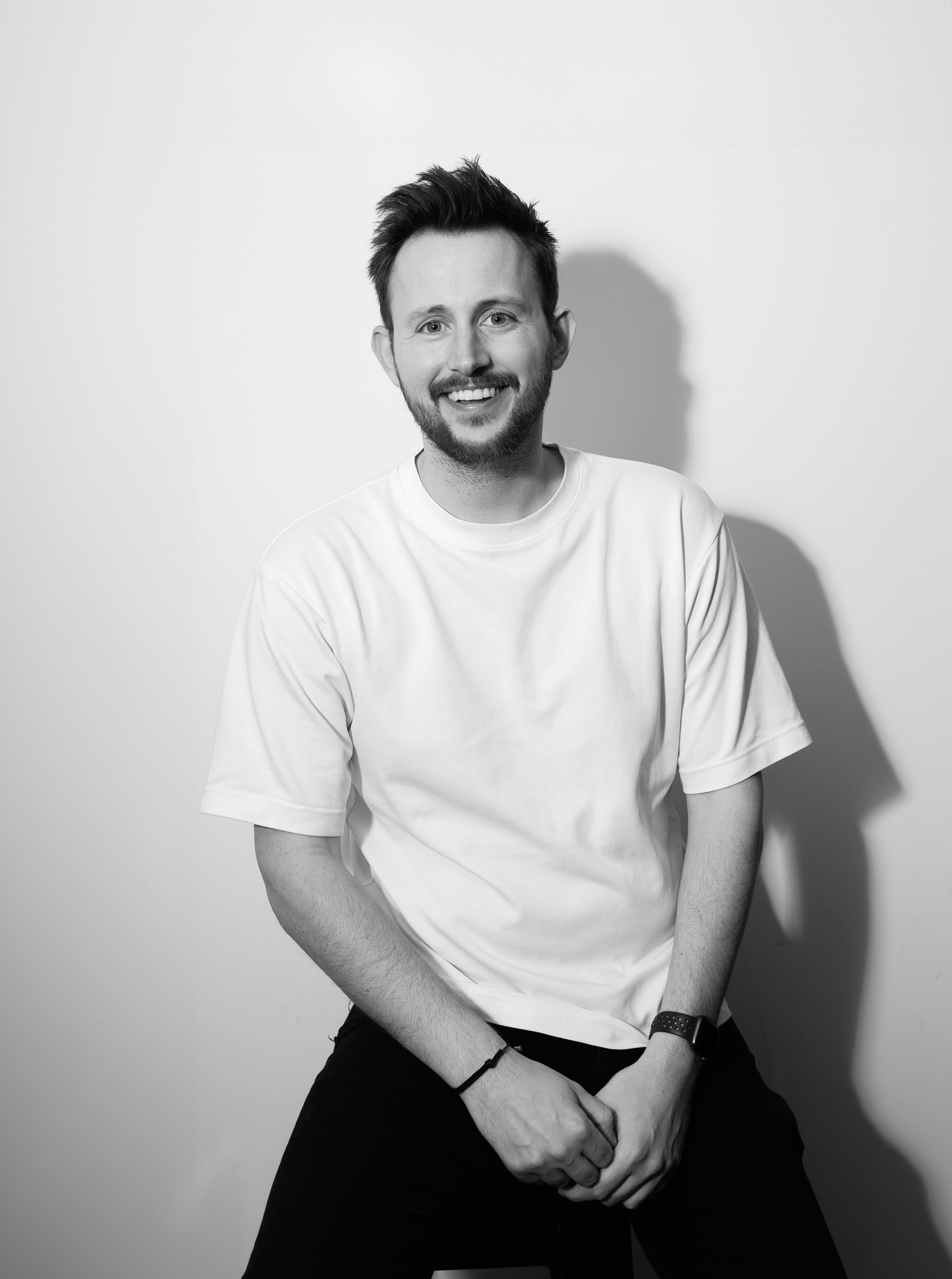 A man in a white T-shirt and dark trousers sits against a plain wall, with his shadow cast to the right.
