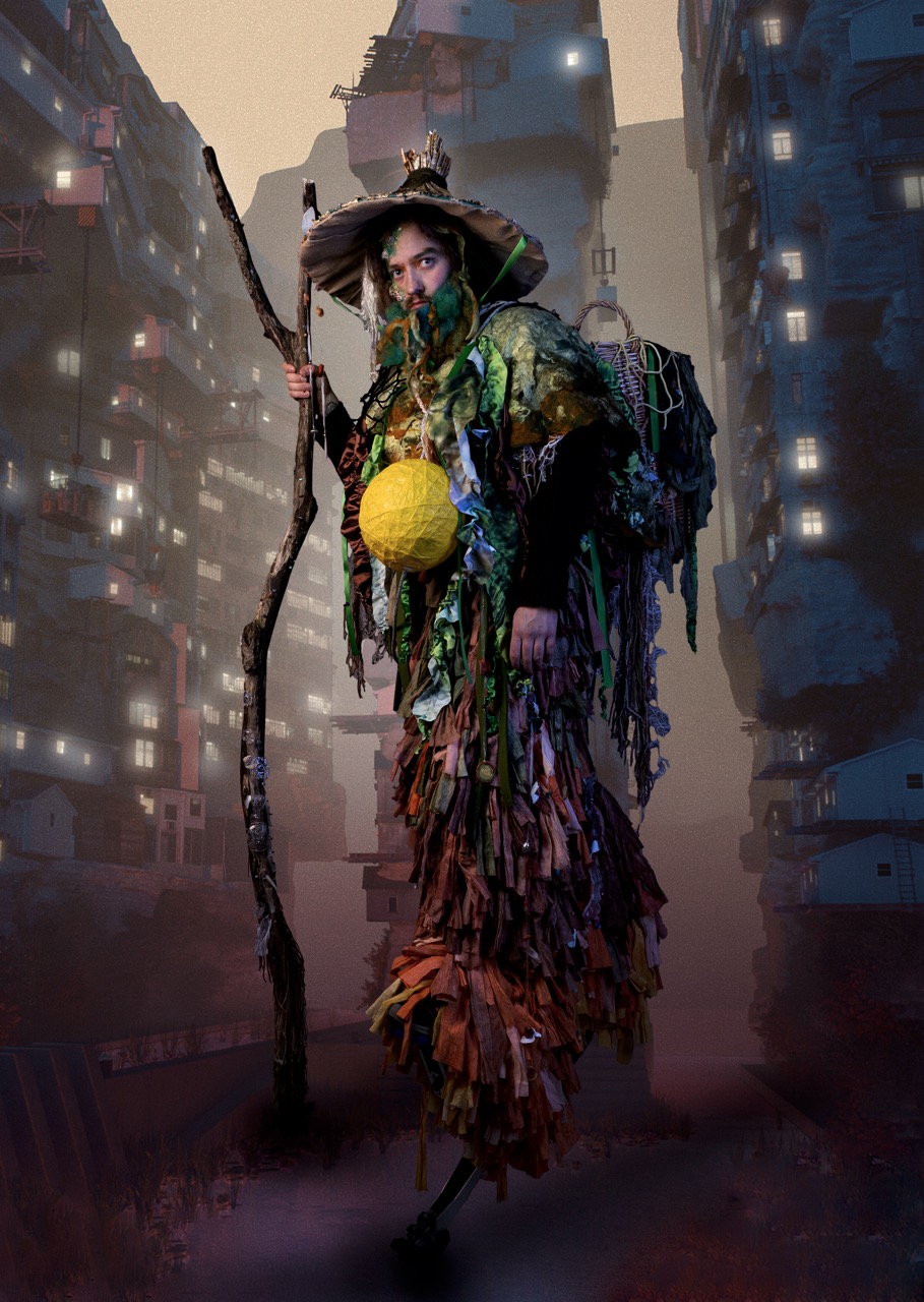 A person in a tall, multicoloured costume with a wide-brimmed hat stands holding a long staff, with towering city buildings fading into mist in the background.