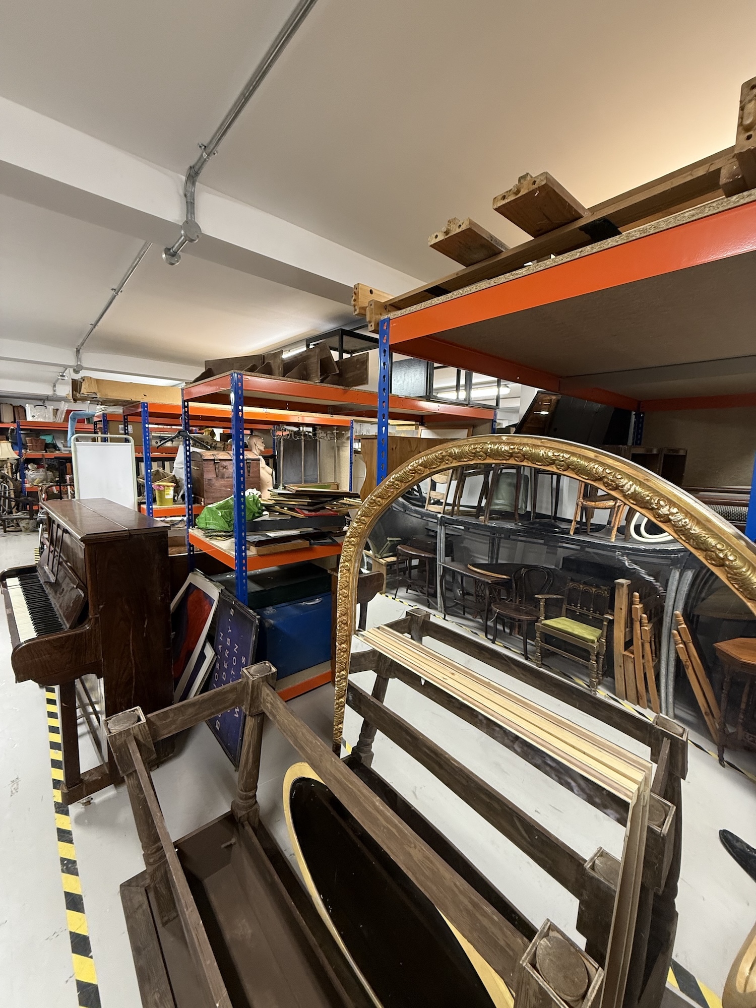 Storage warehouse with blue and orange metal shelving, curved gilt mirror frame in foreground, various furniture and equipment stacked throughout.