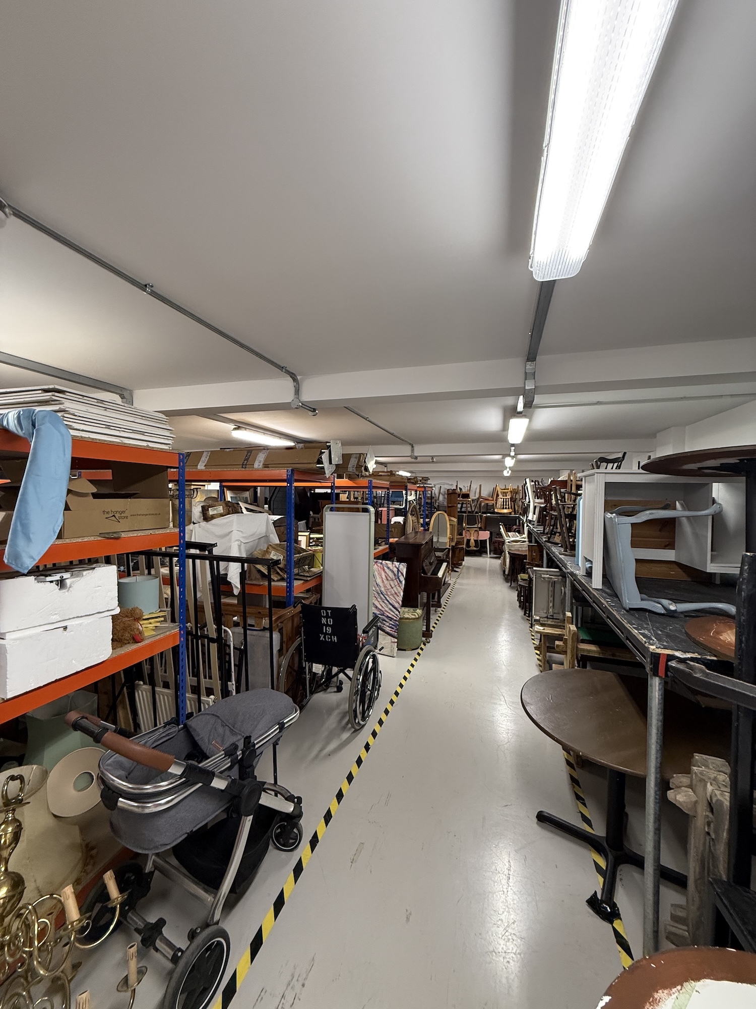 Storage room with fluorescent lighting, blue and orange metal shelving units, and various furniture items arranged along aisles.
