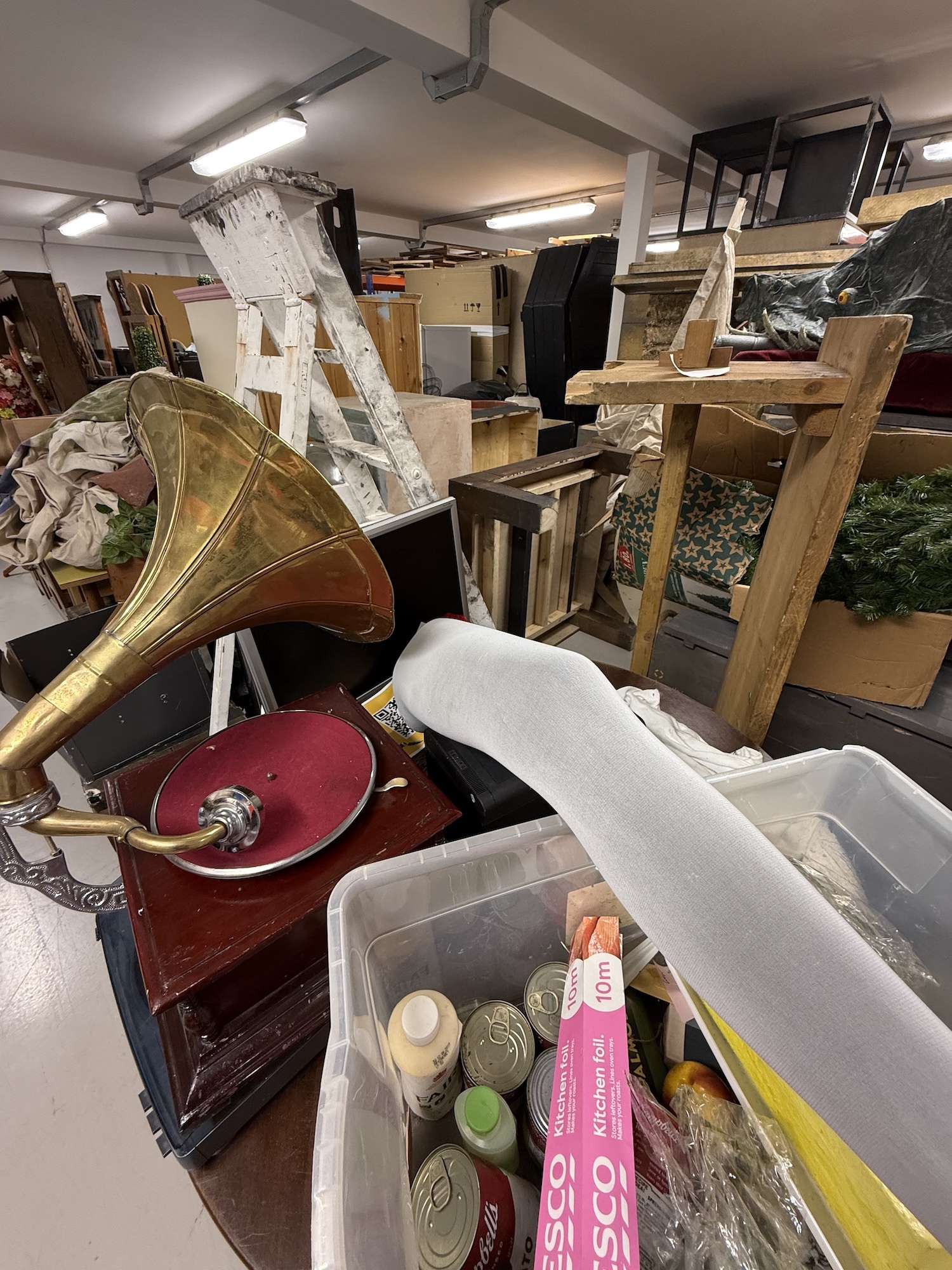 Prop store room area with brass gramophone, wooden furniture, plastic storage boxes, and miscellaneous items under fluorescent lighting.