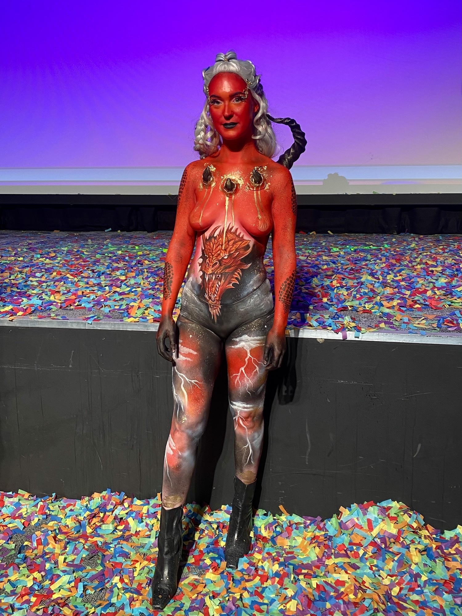 A model with bright coloured body art poses for a photo. Confetti is all over the floor beneath them and on the stage behind them.