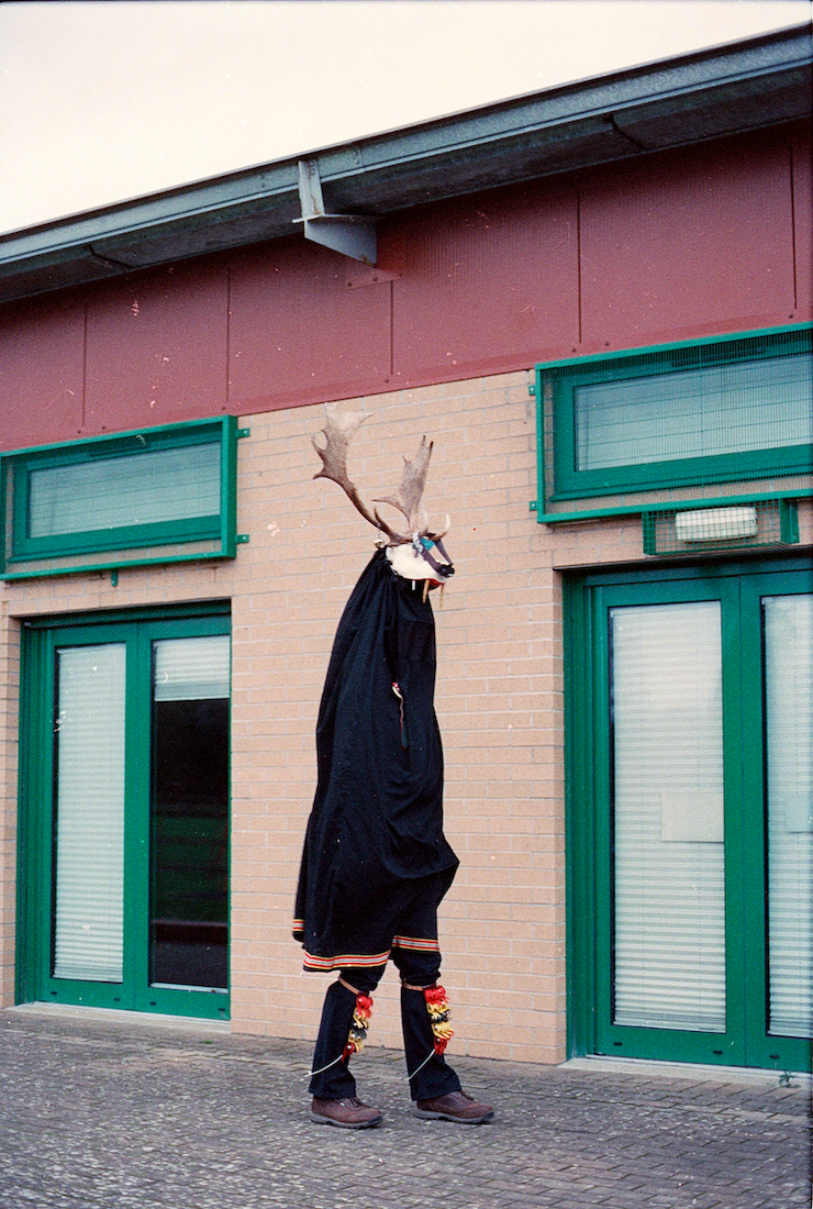 A tall figure wearing a cloak and hood, and a headdress with antlers, standing outside a building with green door frames and blinds.