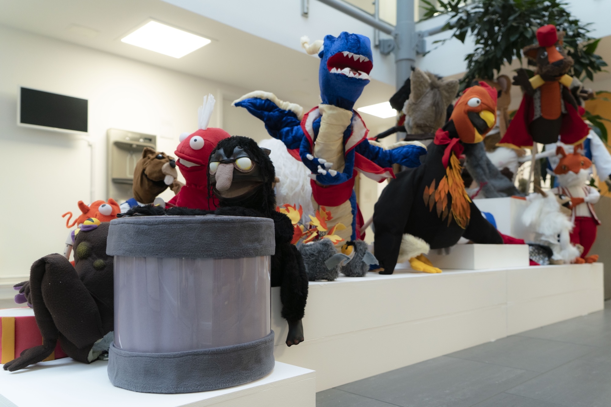 Colourful animal mascot costumes including blue, red, and multicoloured characters sitting on white steps in bright indoor space.