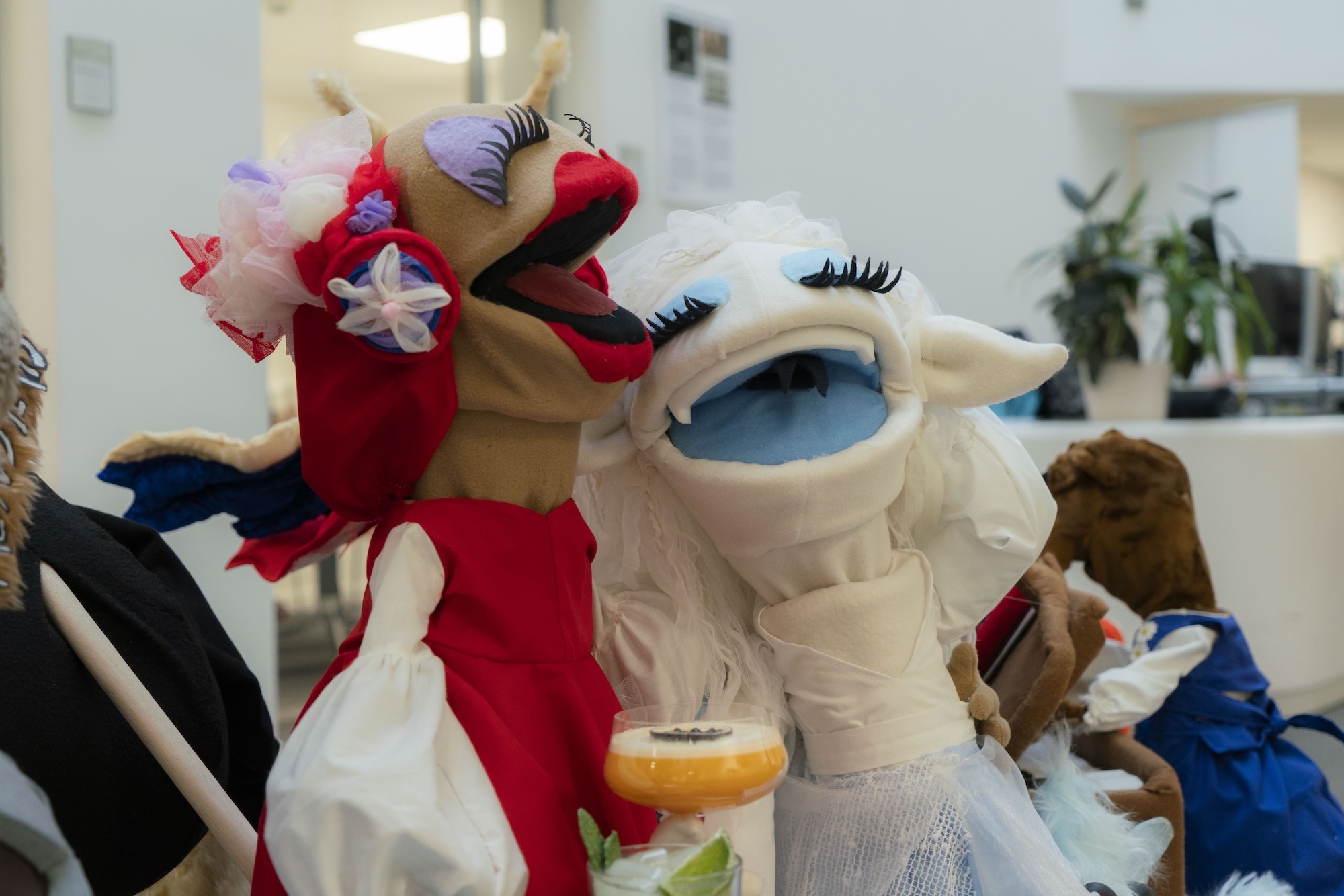 People in colourful animal costumes including red dragon with white flowers, white creature with blue eyes and black eyelashes indoors.