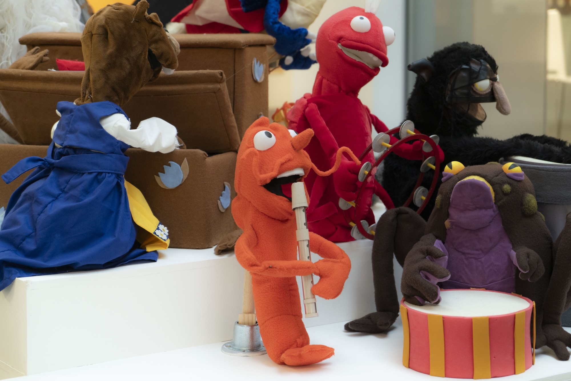 Colourful puppet characters including orange dragon with microphone, blue creature, red dinosaur, and brown animals arranged on white surface.