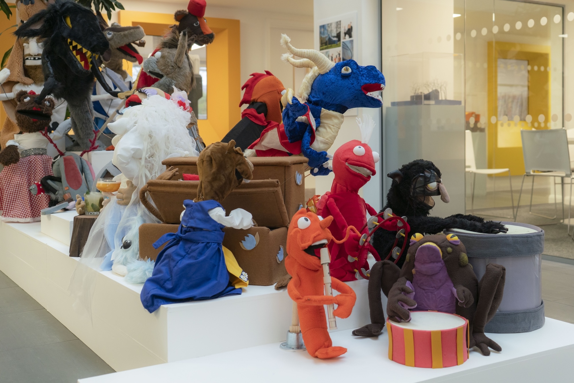 Colourful puppet display with red, blue, orange and brown fabric characters arranged on white platform with wooden chest and striped drum.