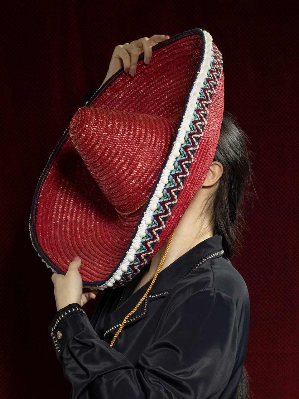 Studio shot of a model obscuring their face with a large, red thrifted sombrero, dark red background.