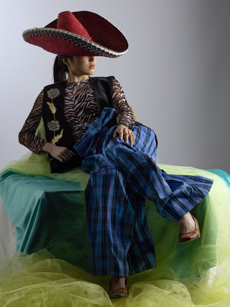 Model wearing a thrifted outfit with a large red sombrero and loose blue trousers, sat on a bed of green and yellow velvet, white background.