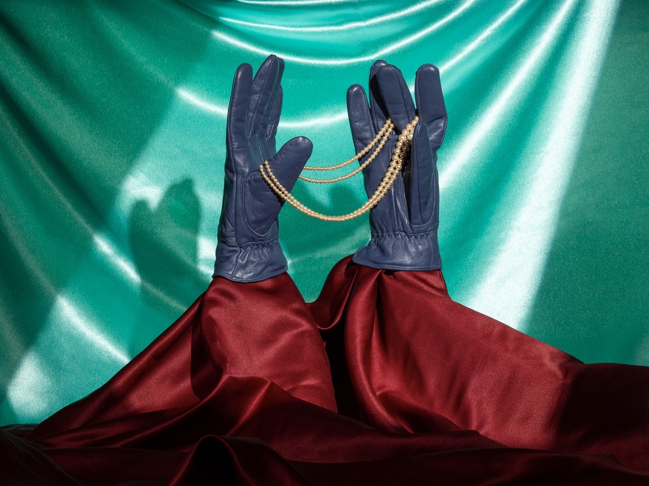 Studio shot of a piece of jewellery being held up by a model wearing dark gloves and a red velvet dress, silky turquoise background.