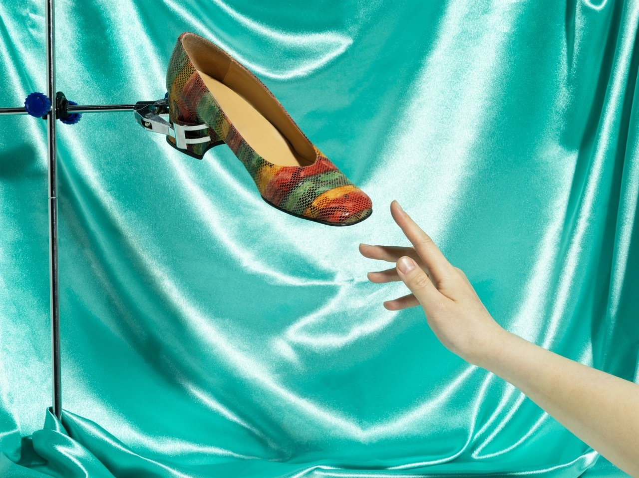 Studio shot of a colourful shoe held on a stand with a model's hand reaching out to touch it, silk turquoise background.