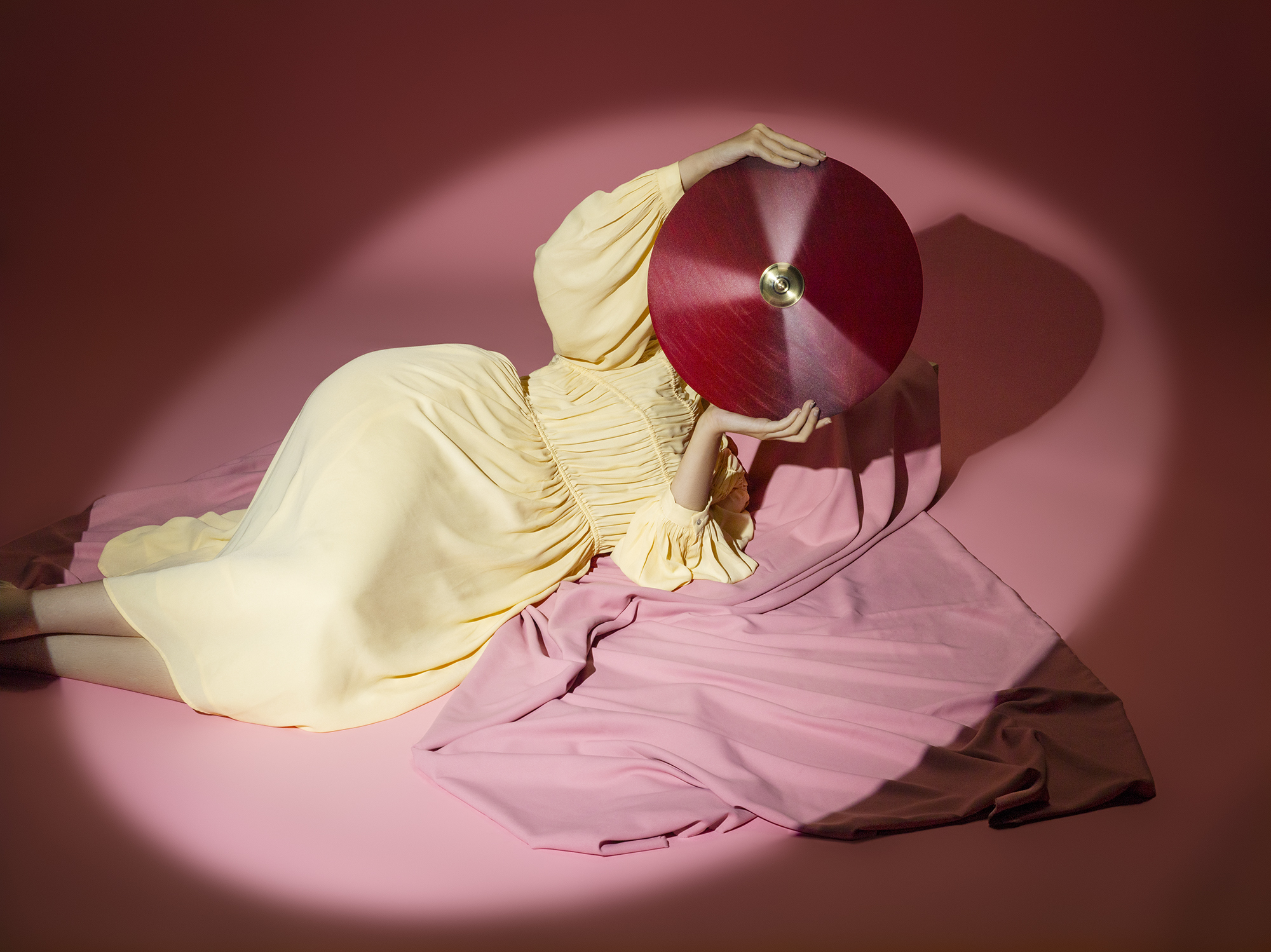 The model is lying down on the floor, hiding her face behind a red lampshade in the spotlight