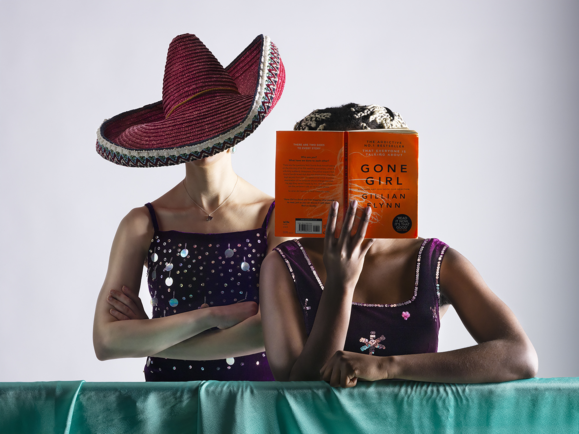 Uncanny Fashion photoshoot featuring two models wearing a hat and holding an orange book, both covering their faces, leaning on a teal surface, white background.