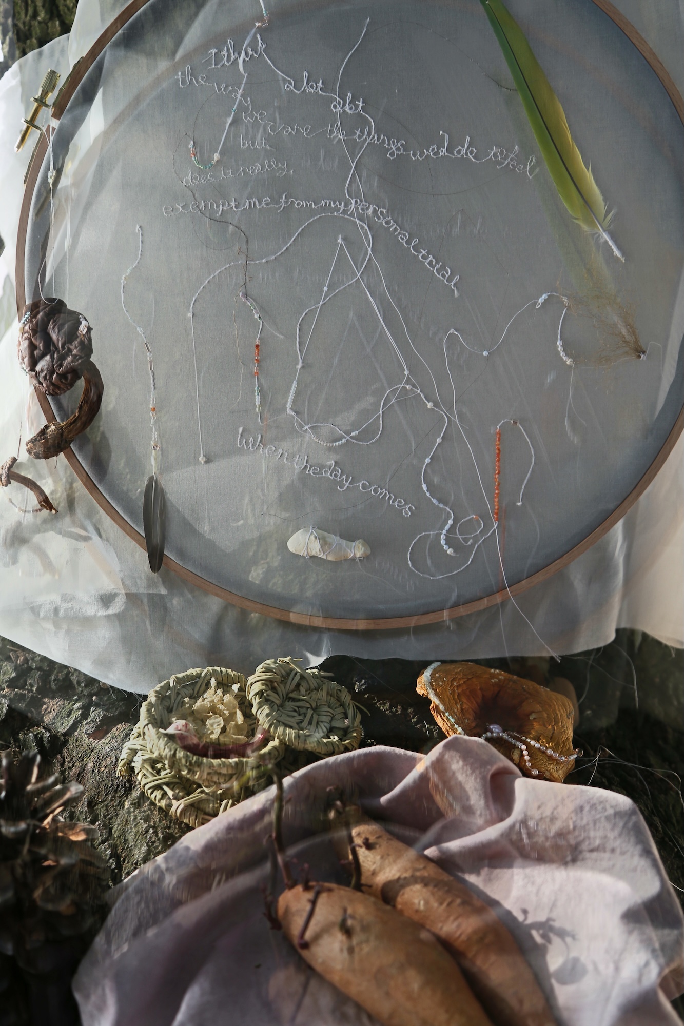A round embroidery hoop with a sheer fabric surface and white stitched outlines hangs above a display of woven baskets and dried objects.