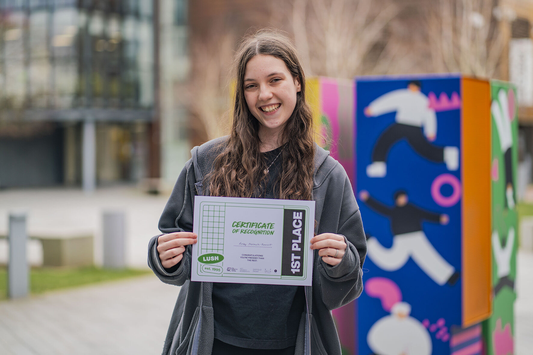 BA (Hons) Graphic Design student R Melmoth-Bennett, holding a certificate for first place, posing for a photo in the courtyard outside University House, Arts University Bournemouth.