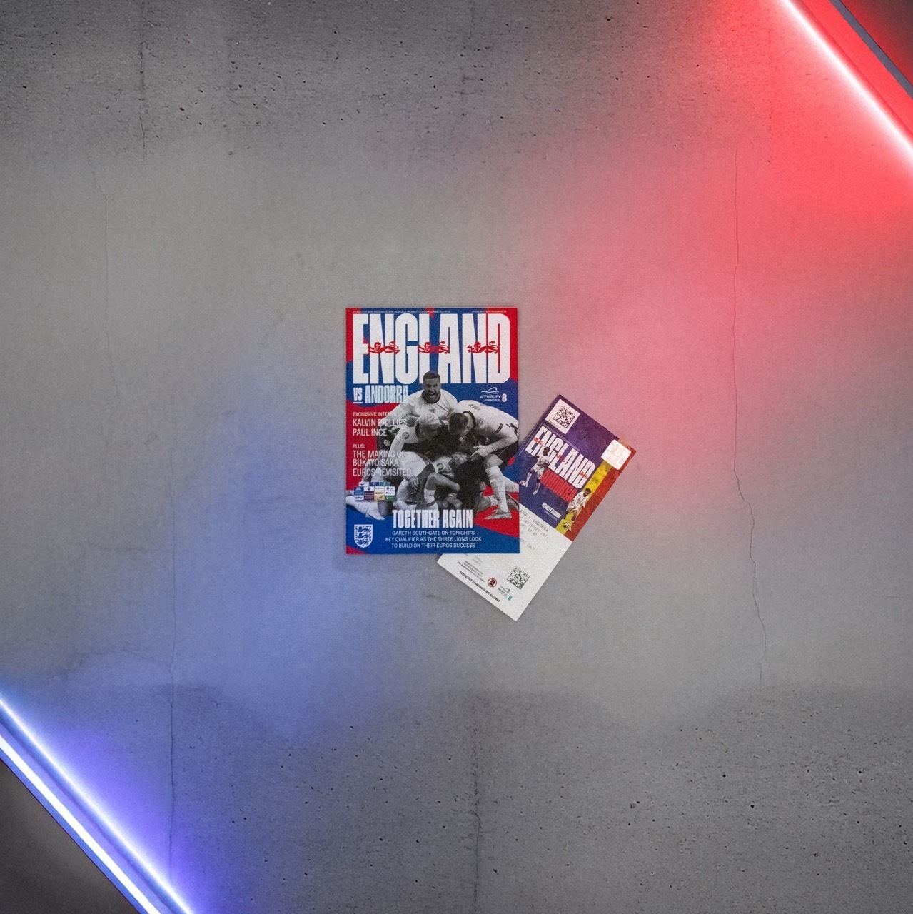 Two England football posters, blue and red, on a concrete wall with blue and red neon lighting illuminating it from each corner diagonally.