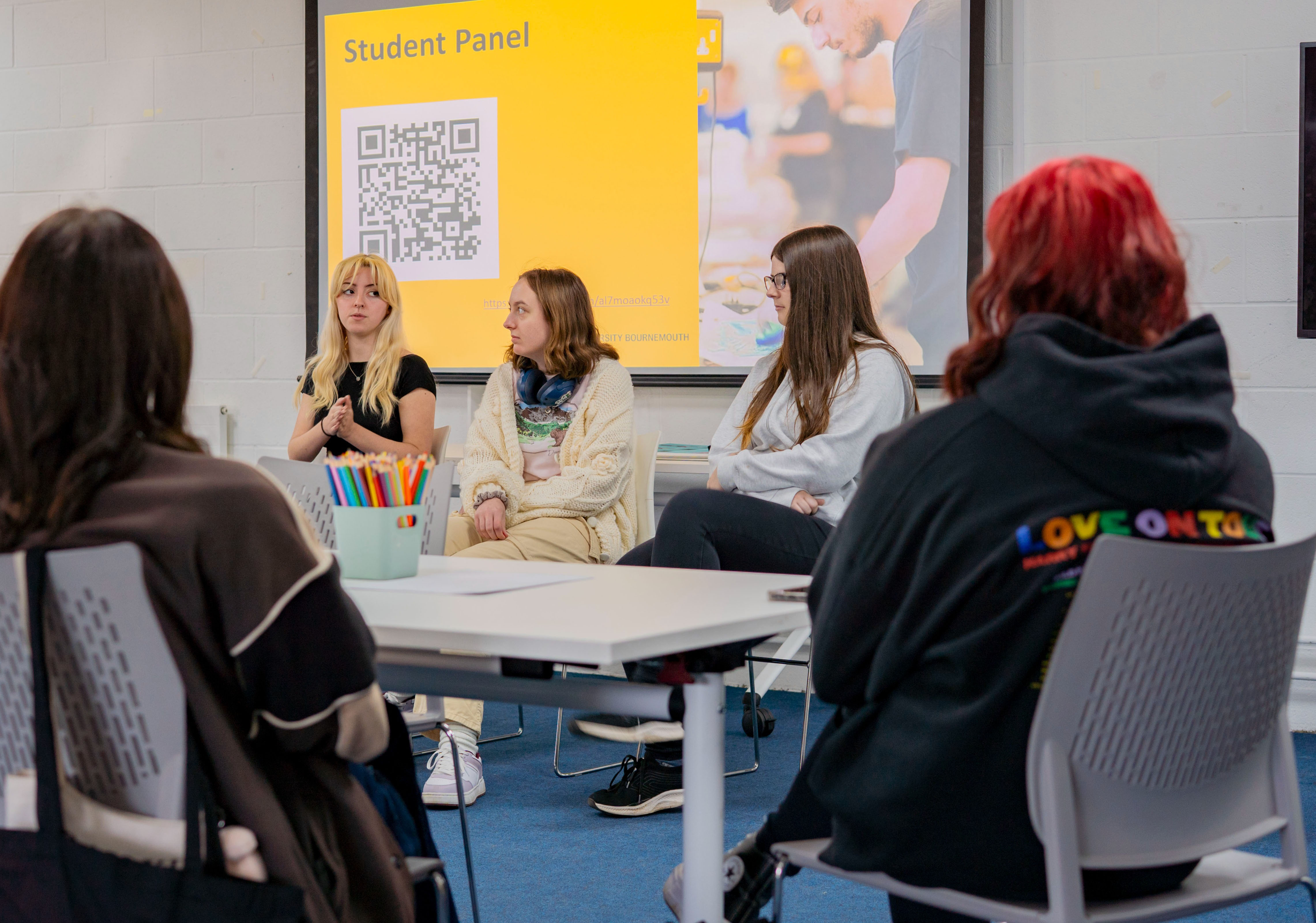 Students seated at white table during panel discussion, yellow "Student Panel" banner with QR code visible in background.