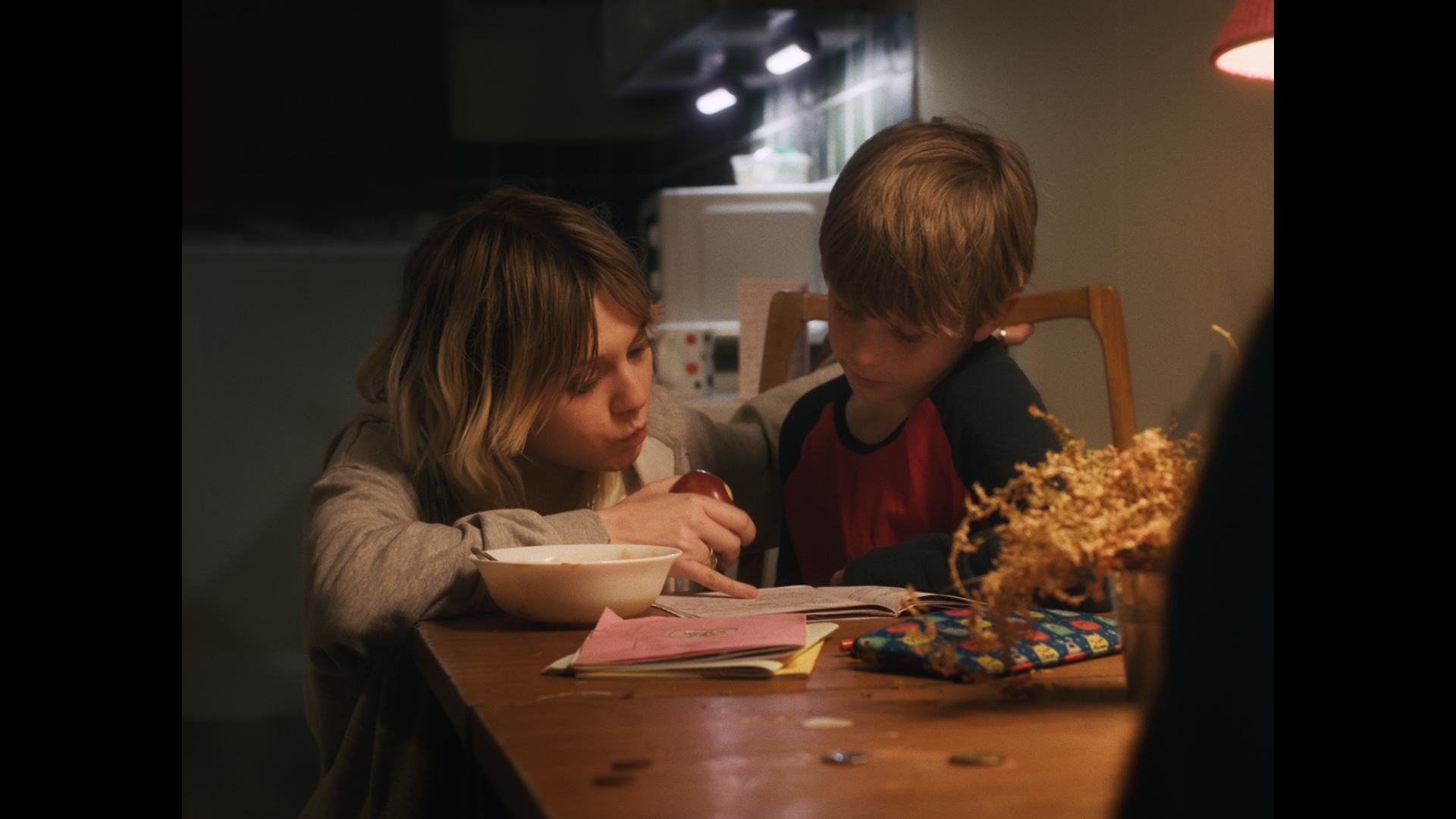 A girl with shoulder-length hair, wearing a grey sweater, leans over a table to guide a boy in a red and black shirt, who is looking at a book. A bowl and various papers are on the table, with a softly lit room in the background.