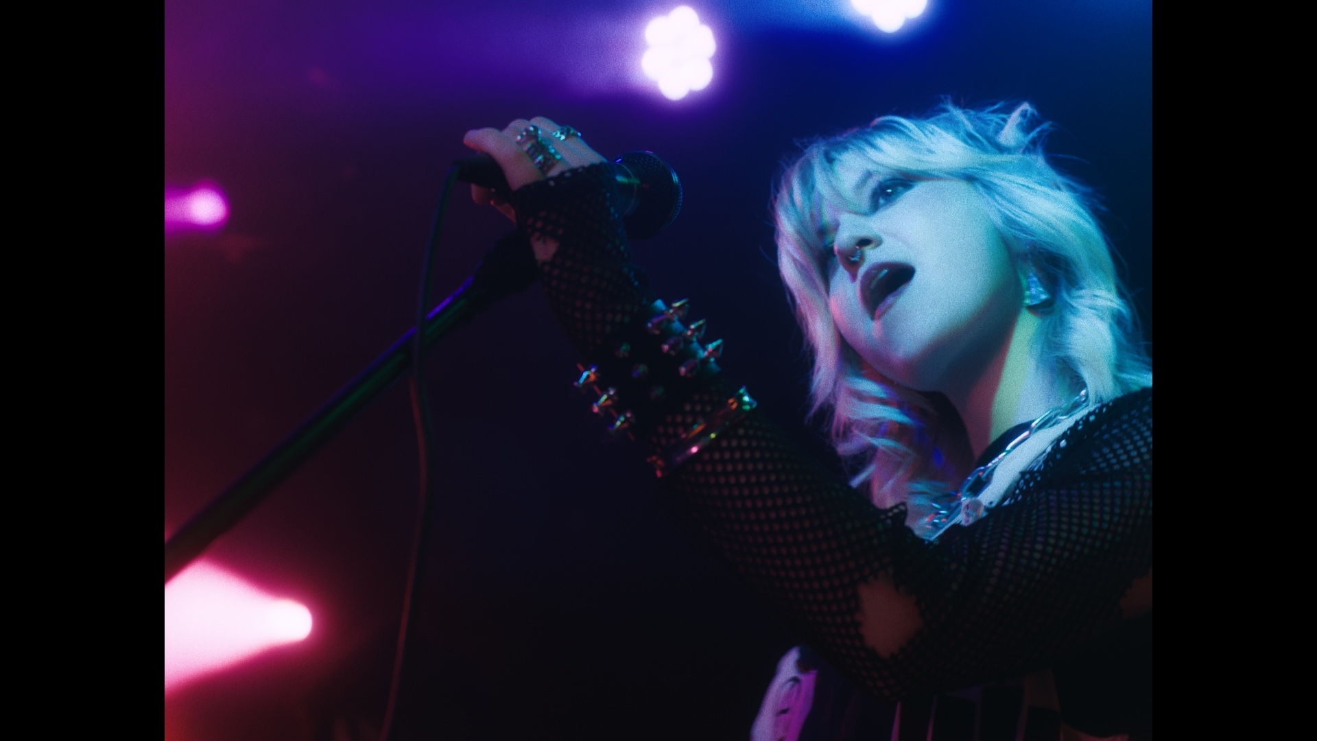 A performer with short, light-coloured hair holds a microphone while singing on stage. They are wearing a black mesh top with spikes, accompanied by colourful stage lighting in pink and blue hues.