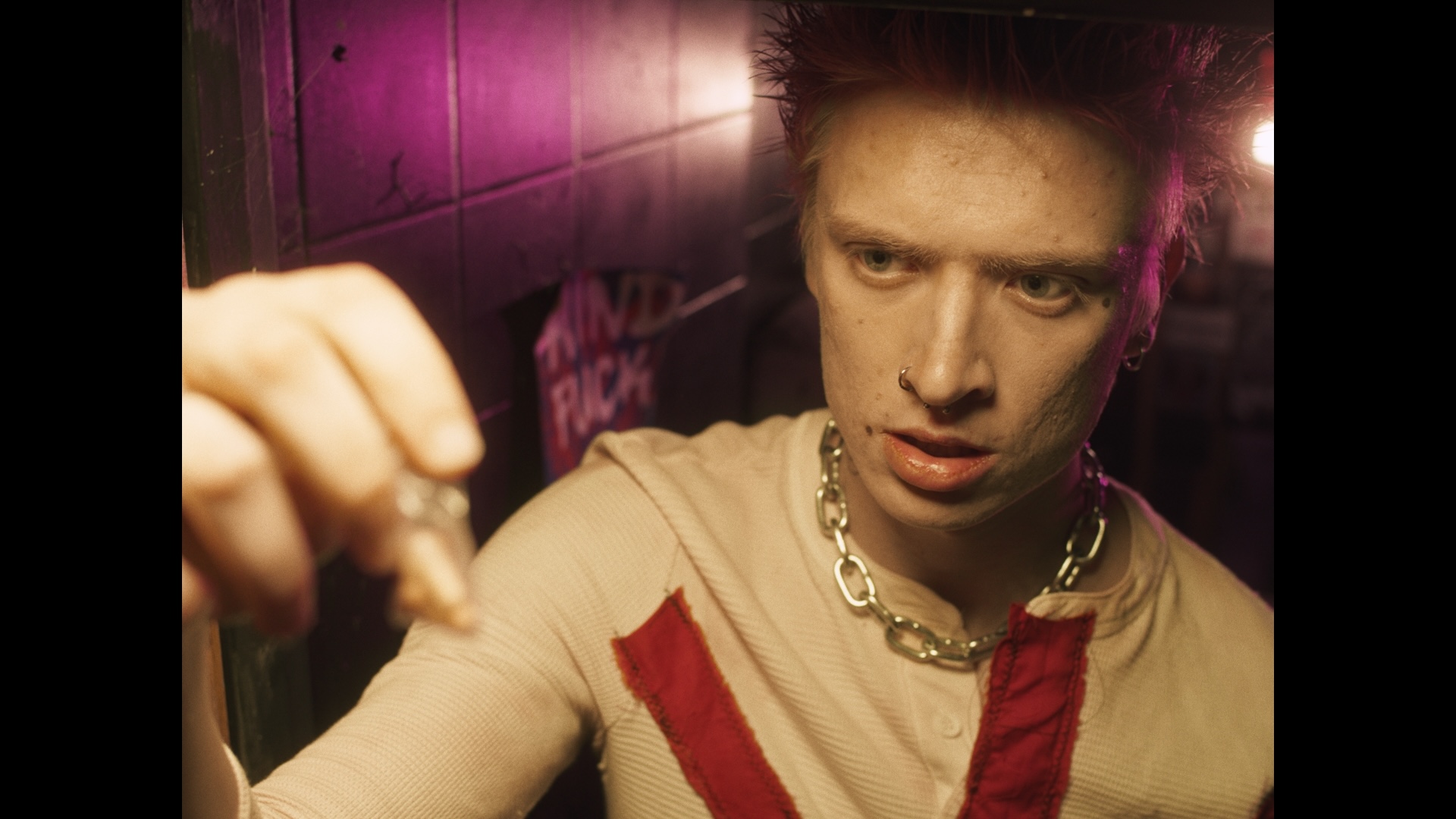 A young individual with short, spiky hair examines a small object held in their hand. The background features dim lighting with pink hues and graffiti on the wall.
